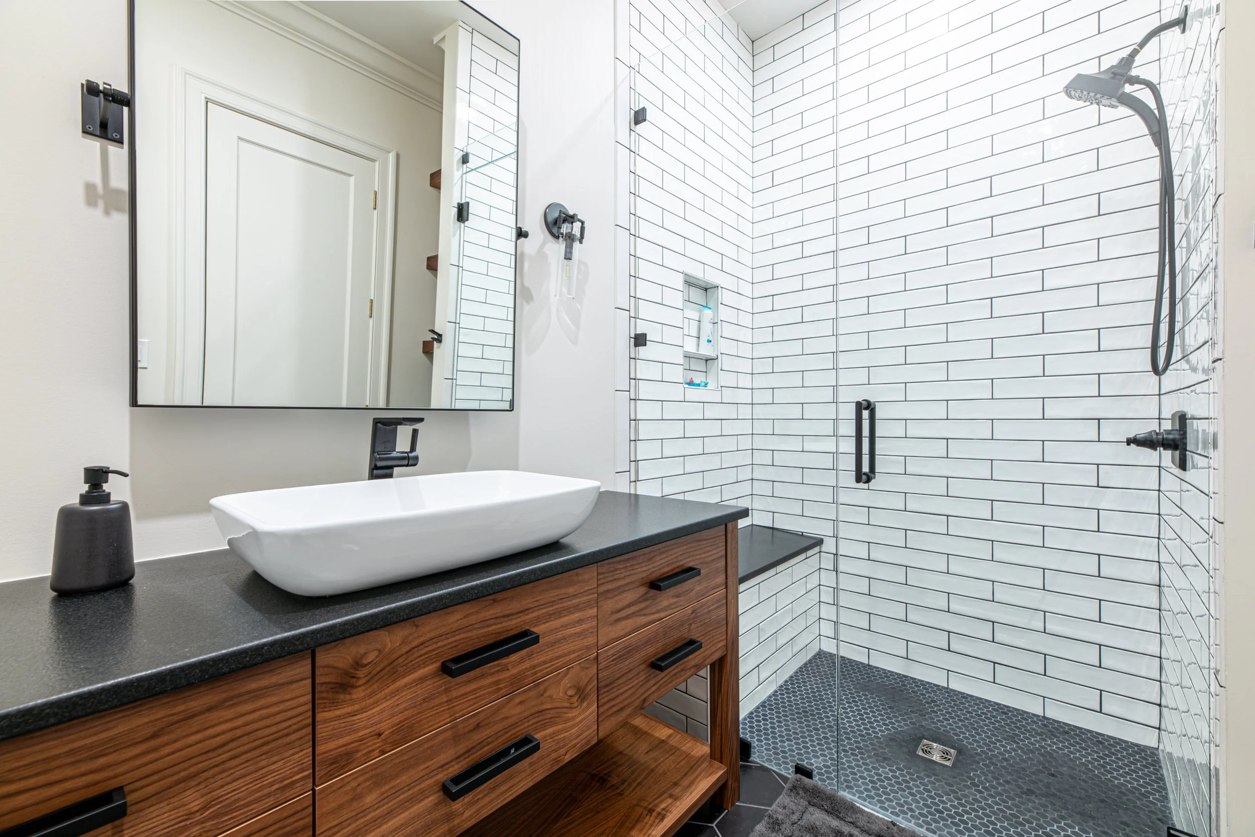 Modern bathroom with wooden vanity, vessel sink, large mirror, and walk-in shower with black fixtures and white subway tiles.