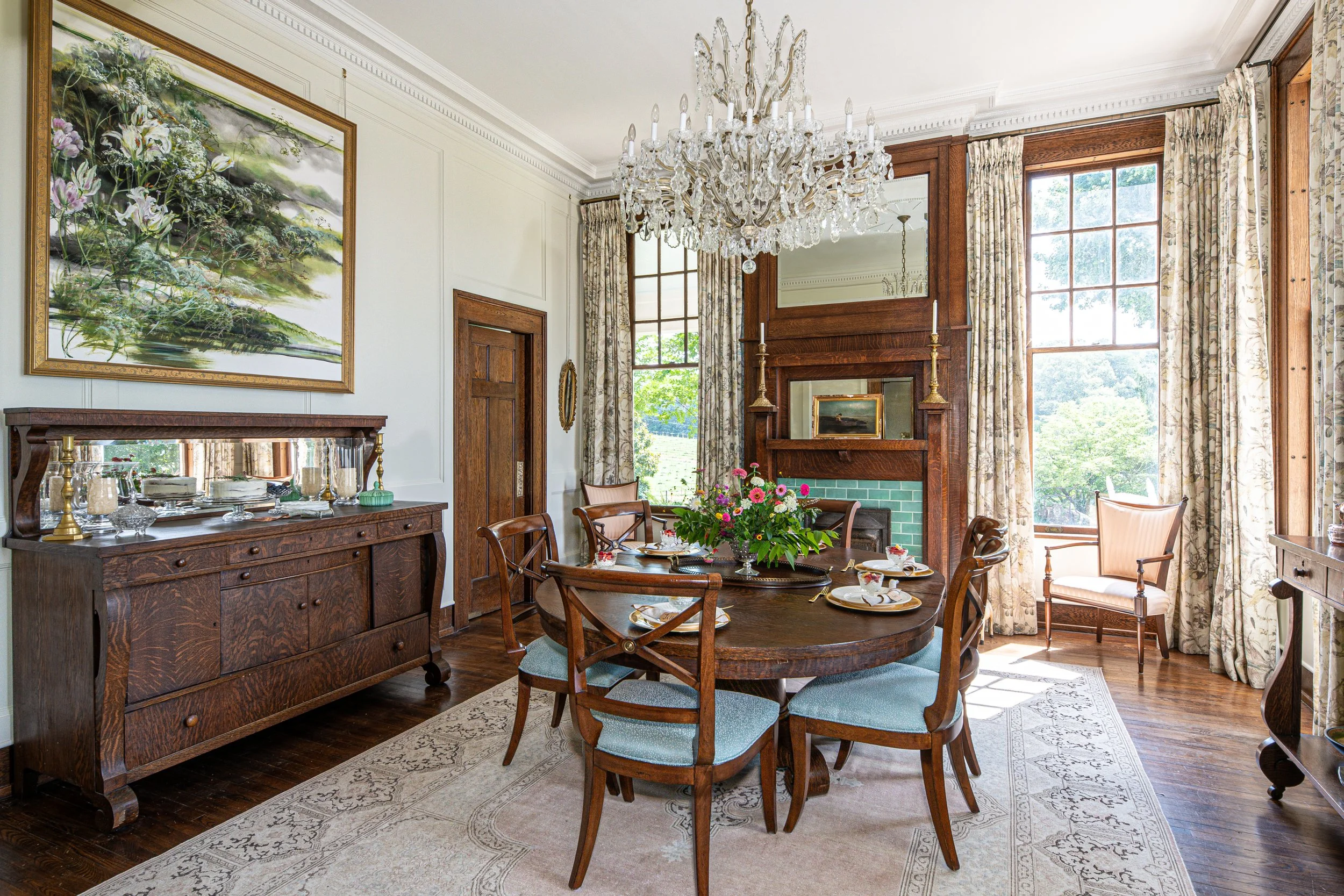 Elegant dining room with a round wooden table set with dishes, a flower arrangement, and six chairs. Features large windows with floral curtains, a crystal chandelier, and dark wood furniture including a sideboard and mantle.