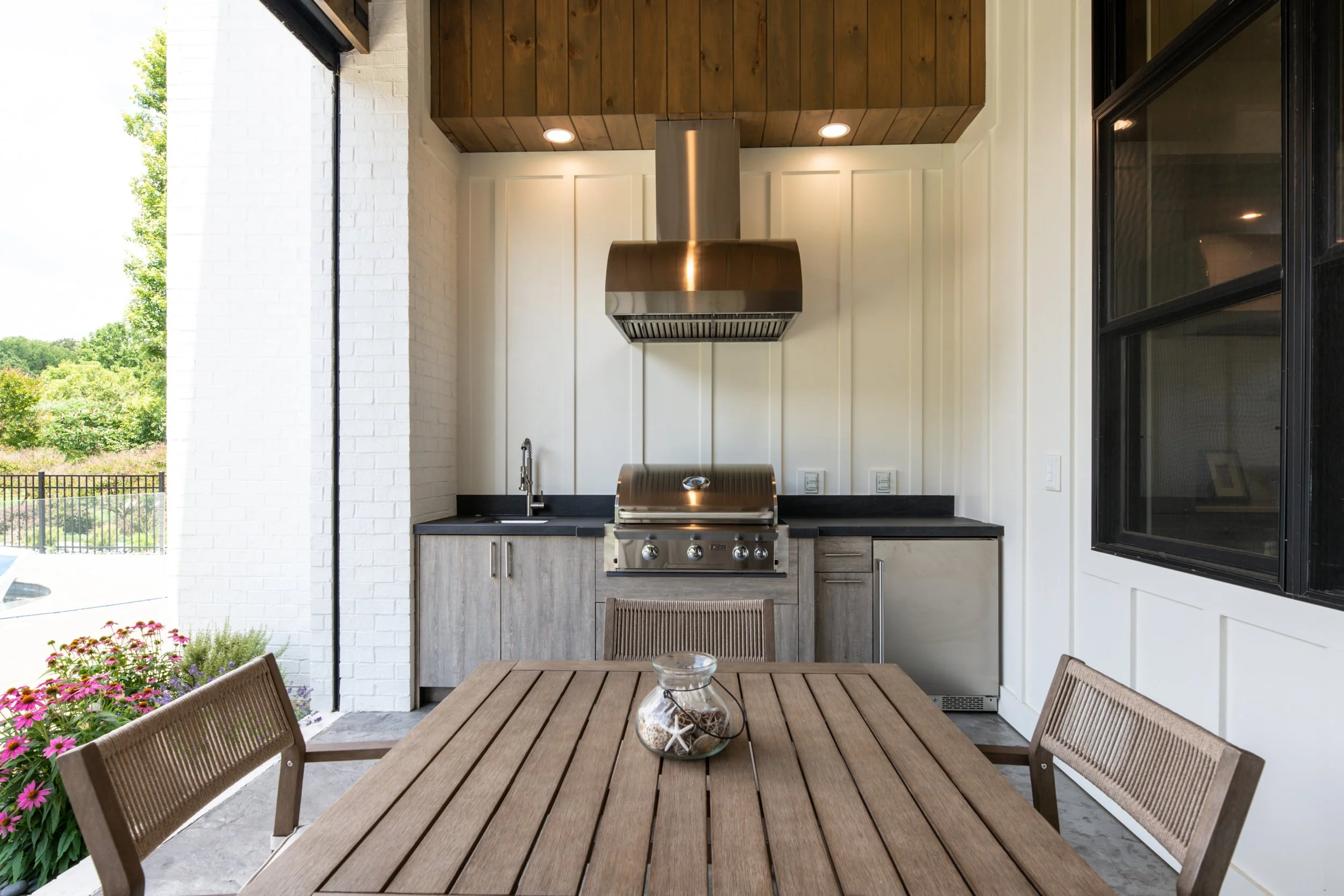 Outdoor kitchen with grill and black countertops, wooden dining table with chairs, flower pot, and large window overlooking green trees