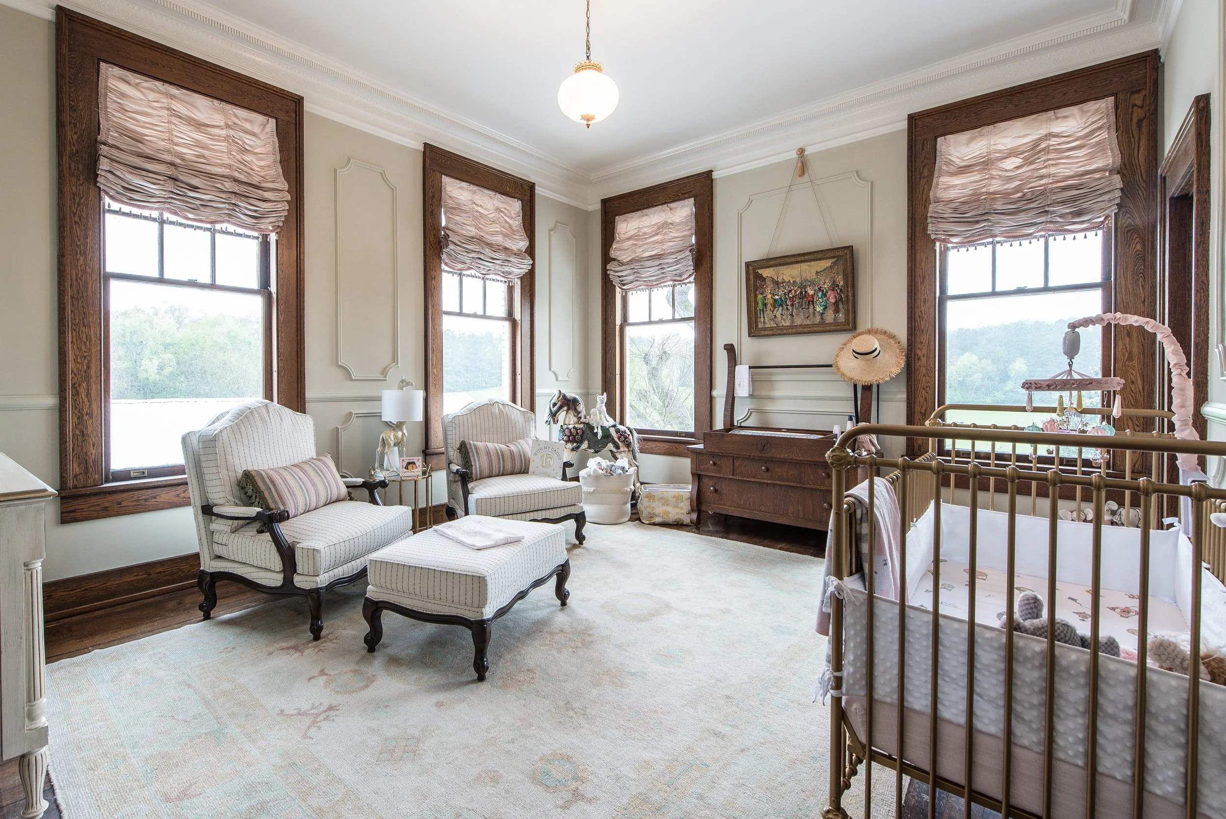 A nursery room with three large windows, draped with pink shades, and furnished with vintage-style white armchairs and a matching ottoman. The room has a wooden dresser, a crib with stuffed animals, toys, and decorative items, and a ceiling light fix