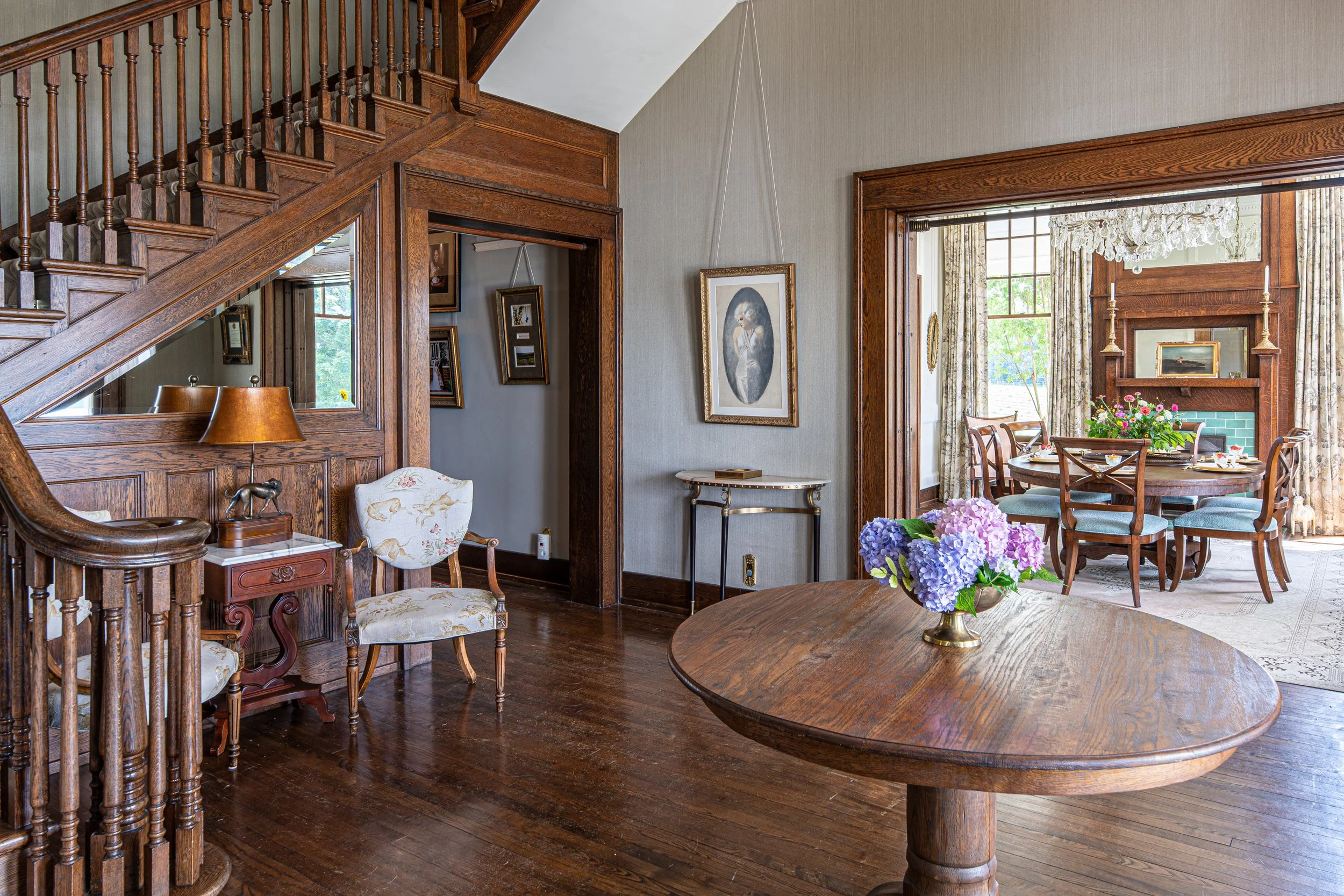 A cozy living room with wood-paneled walls and hardwood flooring. Features a round wooden table with a flower arrangement, a small decorative table with a lamp and elephant figurine, and a staircase with a mirror beneath. An adjacent dining room with