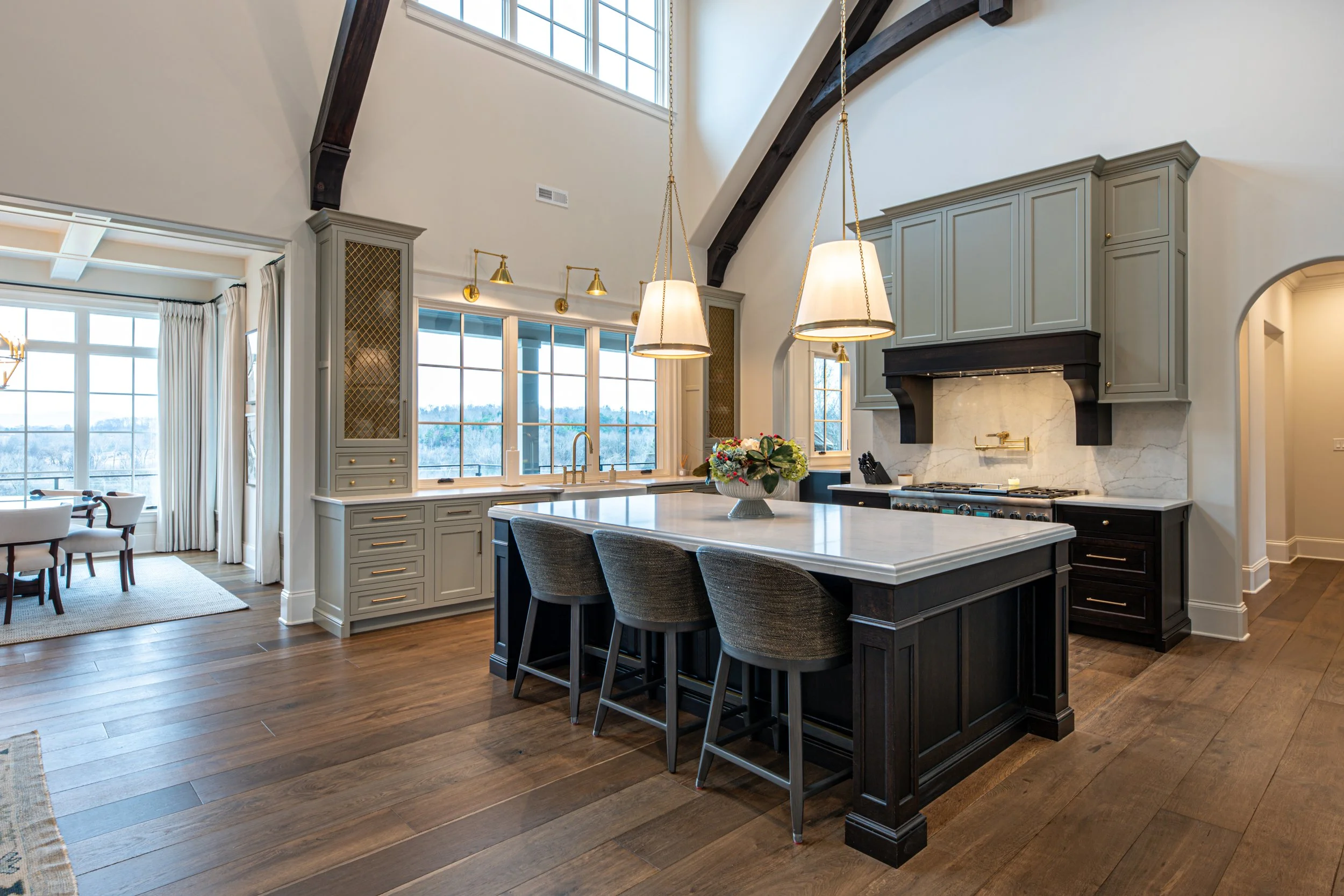 Spacious kitchen with large island, white marble countertops, light gray cabinetry, black accents, wooden flooring, and large windows letting in natural light.