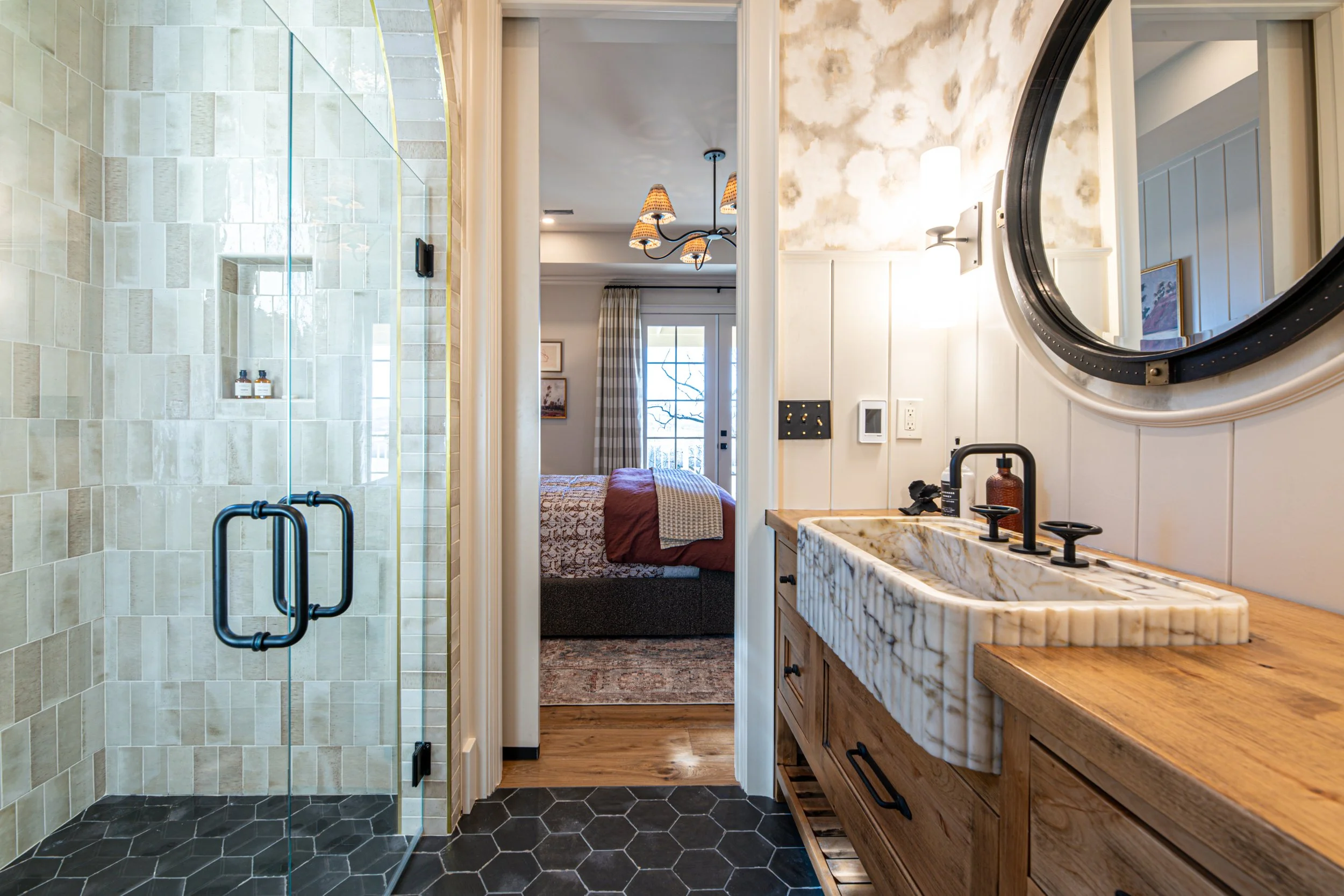 Bathroom with a walk-in shower with glass door on the left, a rustic wooden vanity with a marble sink on the right, and a bedroom visible through the doorway in the background.
