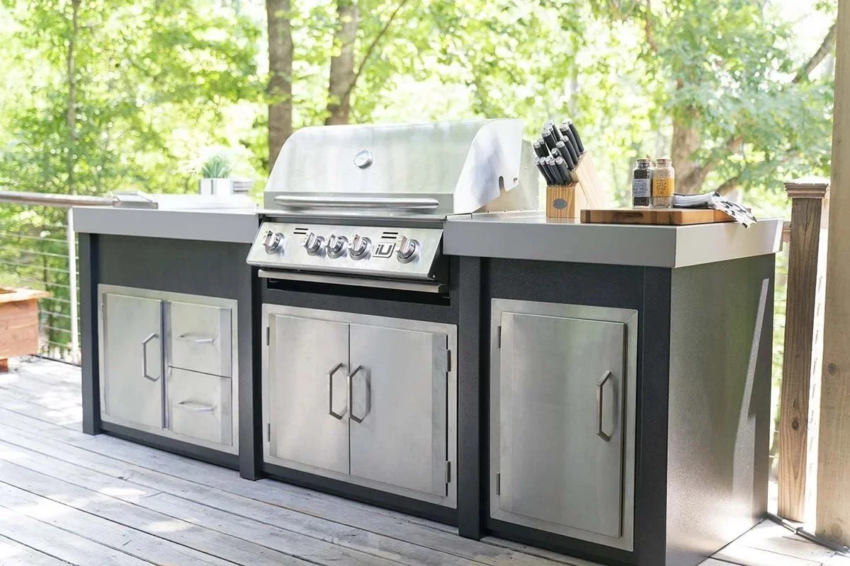 Outdoor kitchen with grilling station and storage cabinets on a wooden deck surrounded by trees.