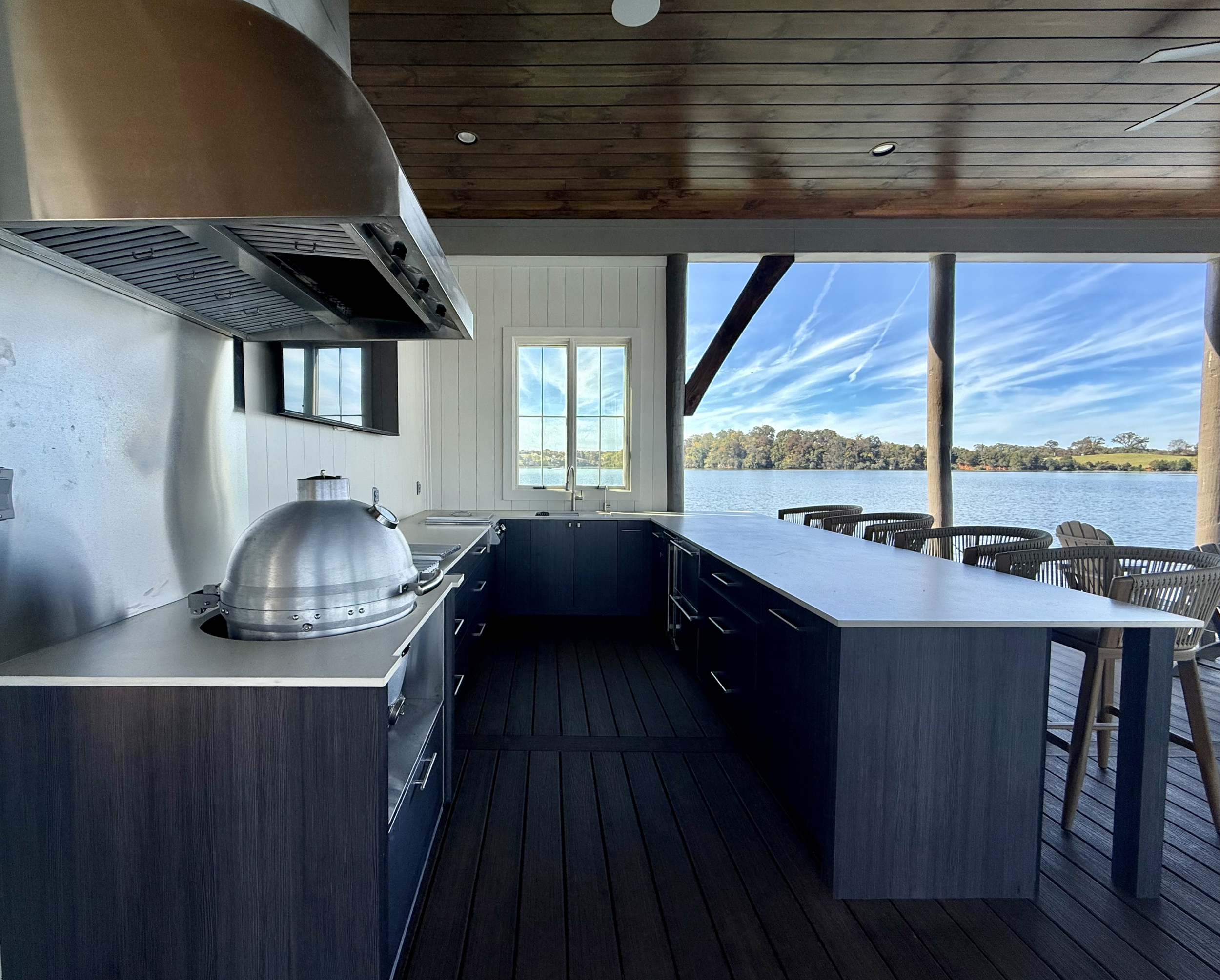Kitchen area with a view of a lake, featuring a long white countertop, dark cabinetry, a window showing the outdoors, and outdoor seating along the water.