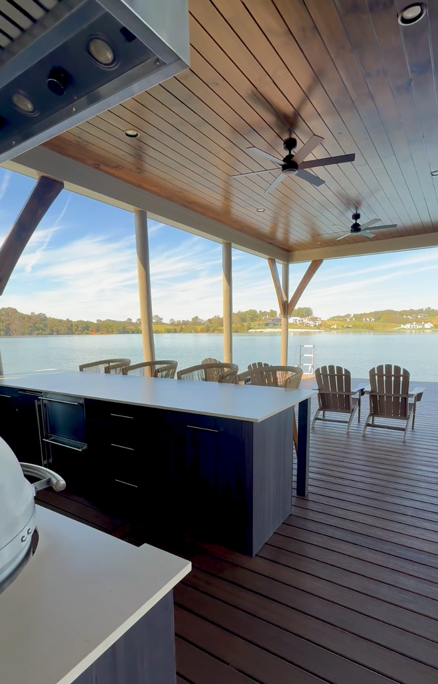 Outdoor covered patio overlooking a lake with wooden deck, ceiling fans, and Adirondack chairs.