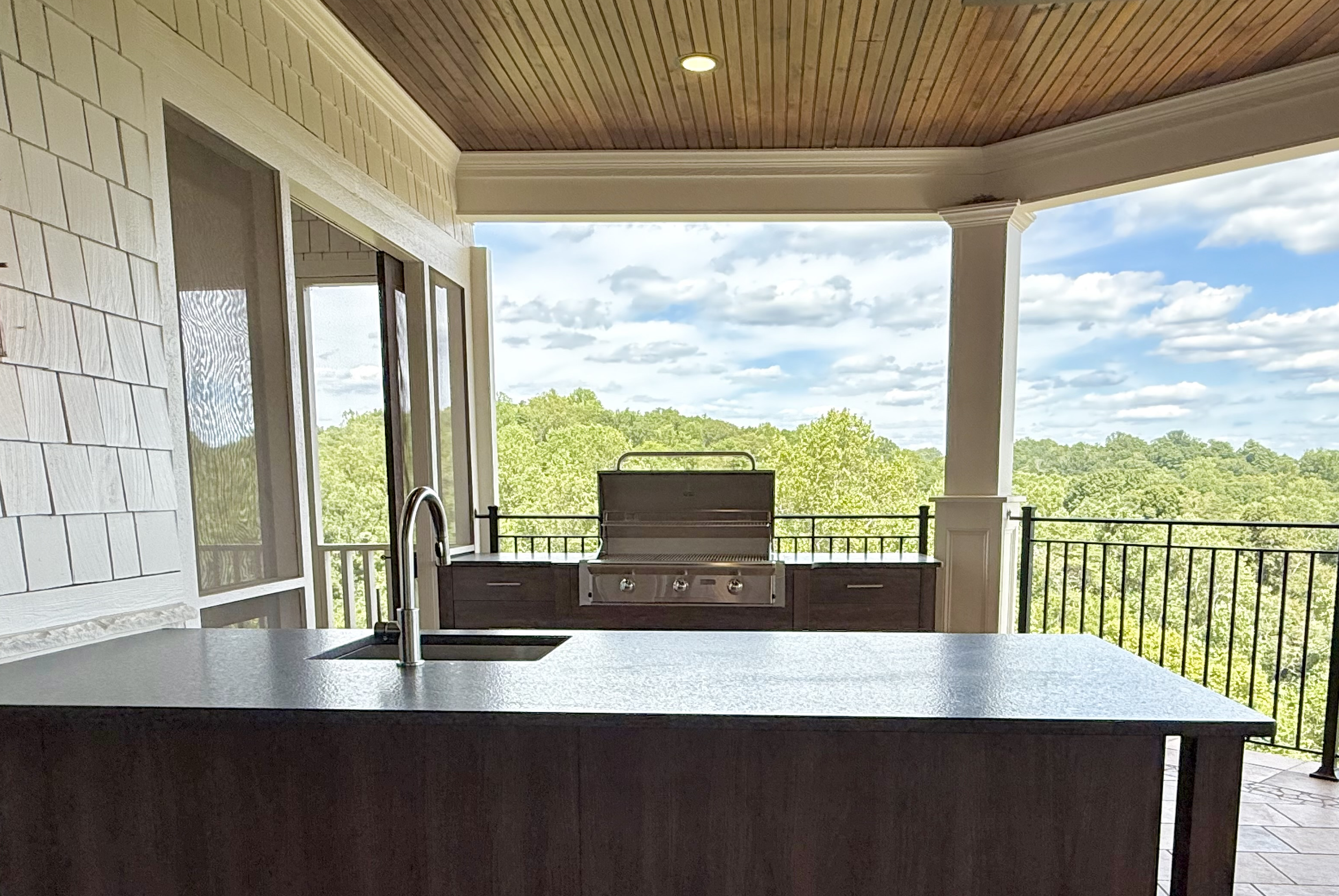 Outdoor kitchen area with a black countertop, a built-in grill, a sink with a modern faucet, and a view of trees and a cloudy sky through a balcony railing.