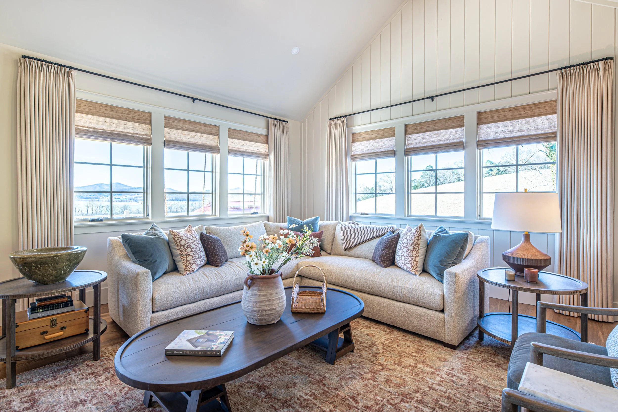 Living room with beige sectional sofa, multiple throw pillows, wooden coffee table with a vase of flowers, side tables with a lamp and decorative items, large windows with brown shades and curtains, and a patterned area rug.