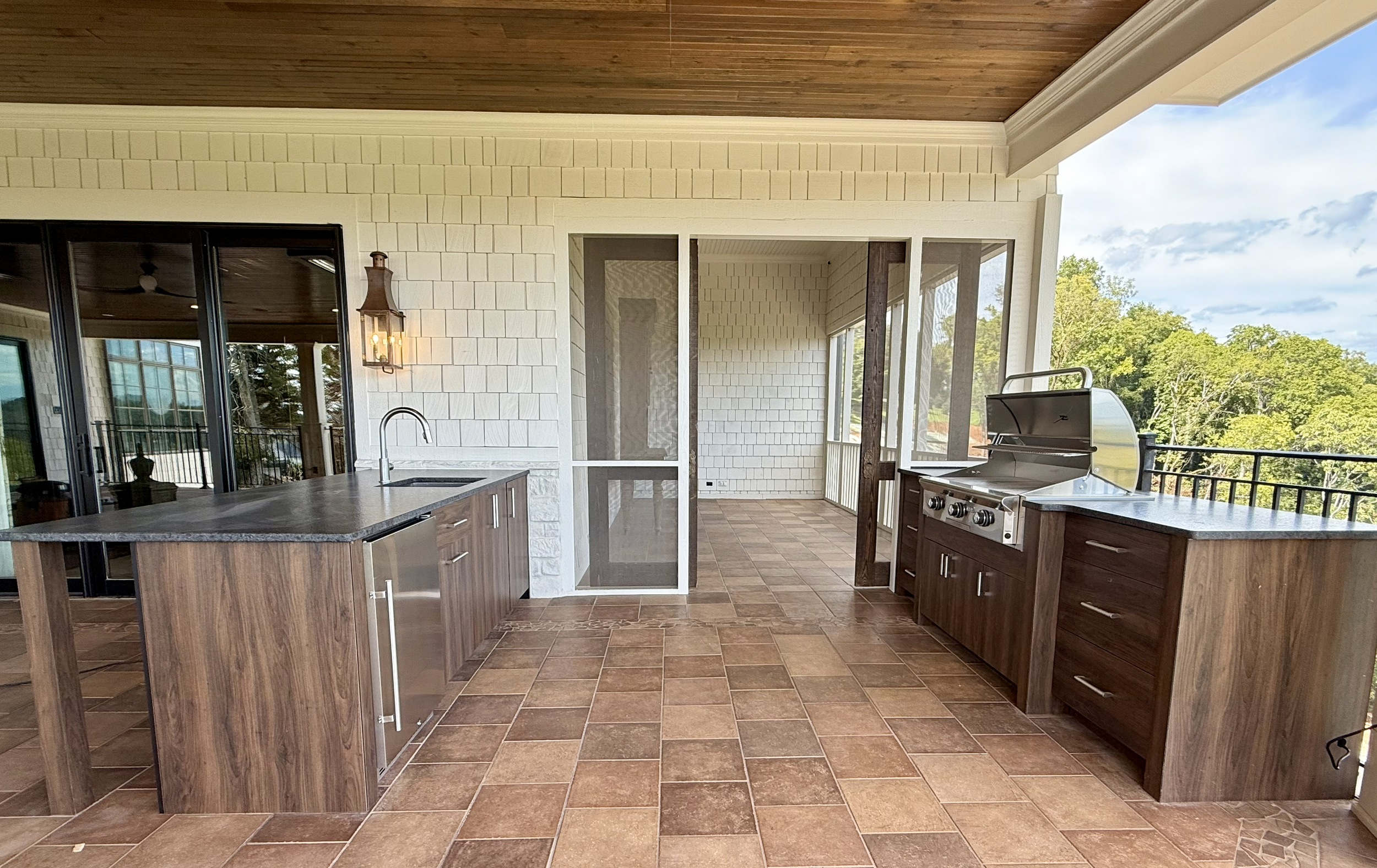Covered outdoor kitchen area with brown tile flooring, wooden cabinets, a barbecue grill, a sink, and a lantern style wall light, overlooking a lush green landscape.