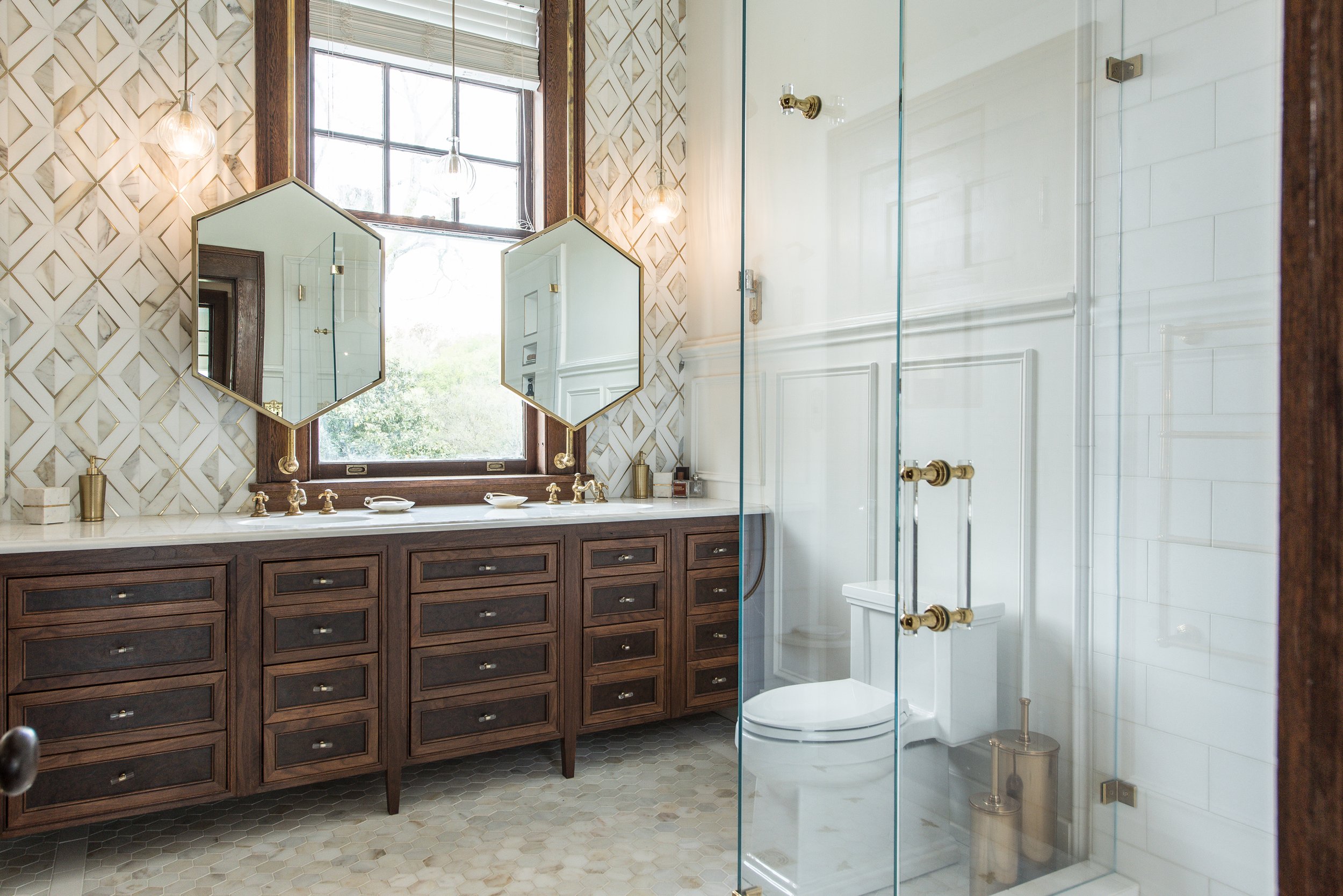 Bathroom with large window, mirror, wooden vanity with multiple drawers, white countertop, and glass shower enclosure with brass fixtures.
