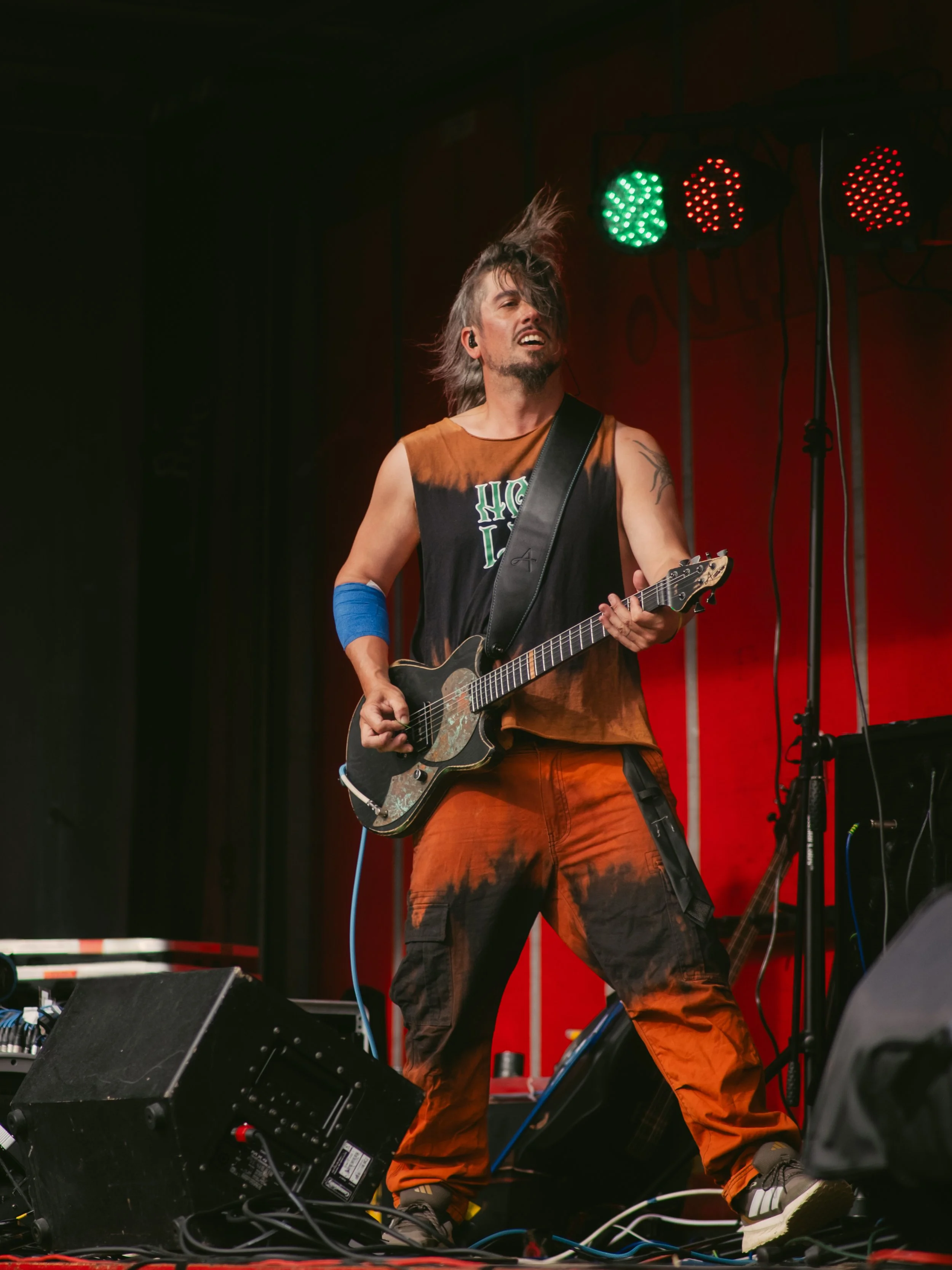 A man with long hair playing an electric guitar on stage, wearing an orange and black sleeveless shirt and orange pants, with colorful stage lights overhead.