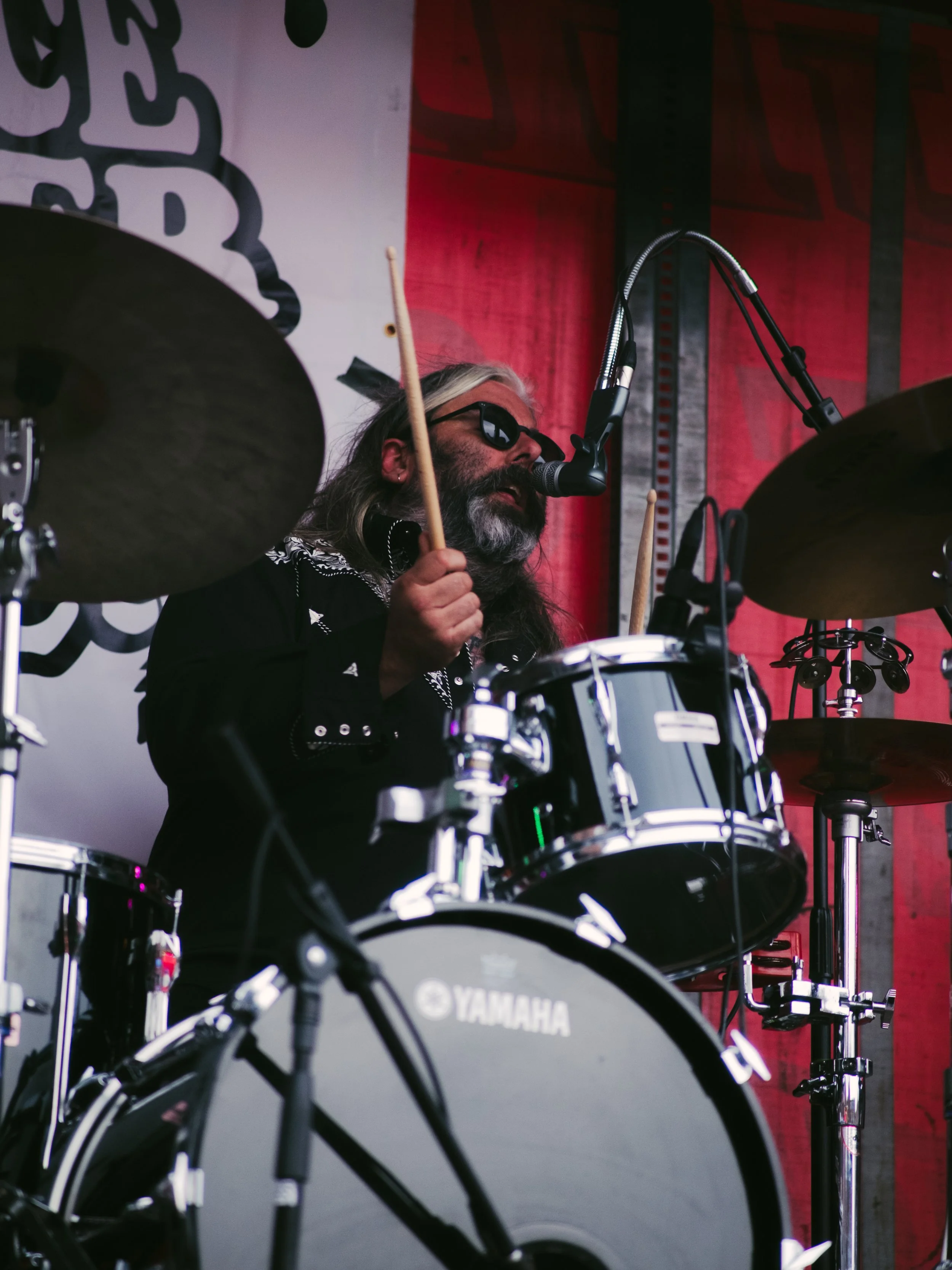 A male musician with long hair, beard, glasses, and black clothing playing a Yamaha drum set on stage, singing into a microphone, with red and gray backdrop.