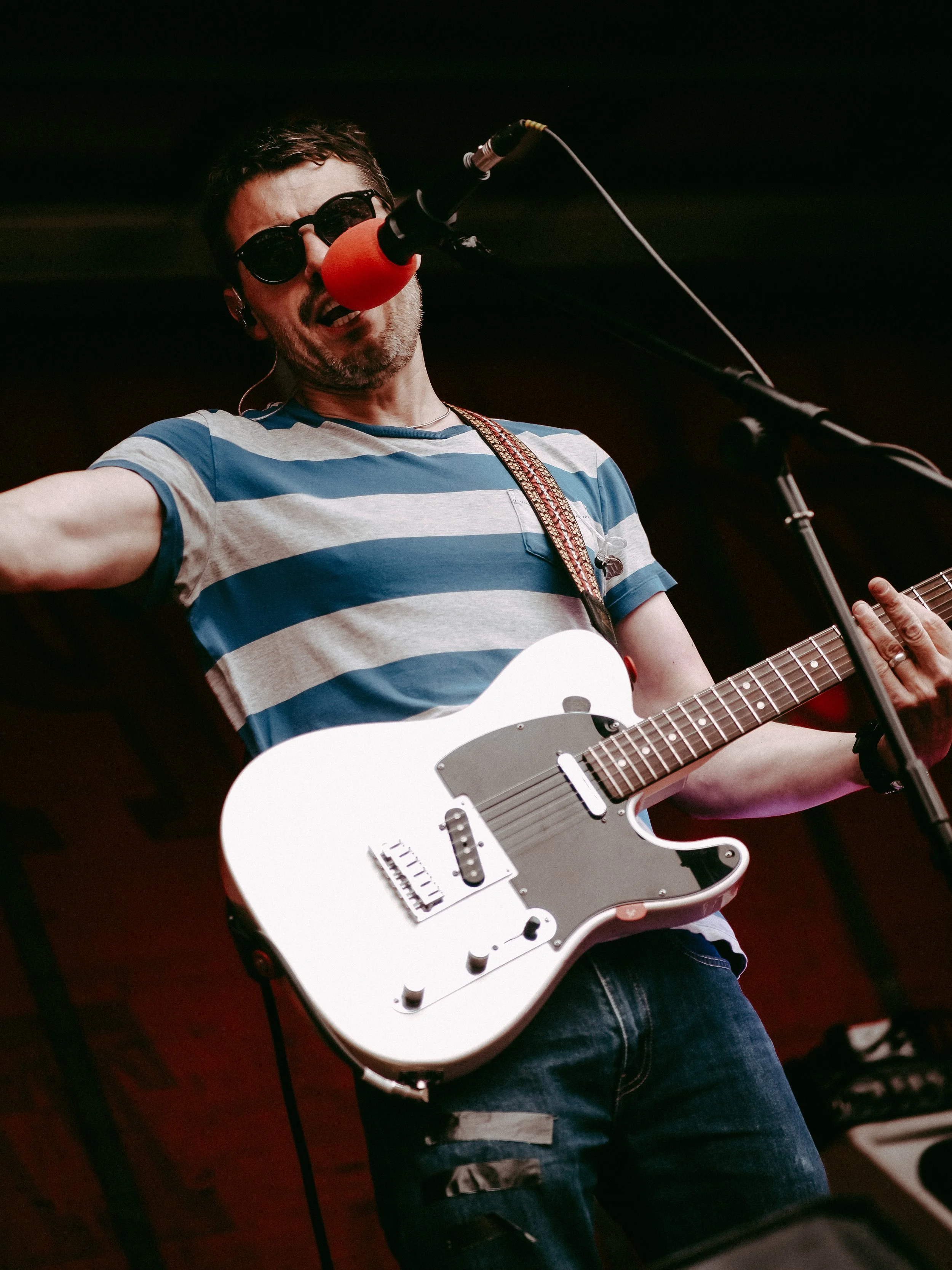 Male musician wearing sunglasses and a blue and white striped shirt playing a white electric guitar and singing into a red microphone.
