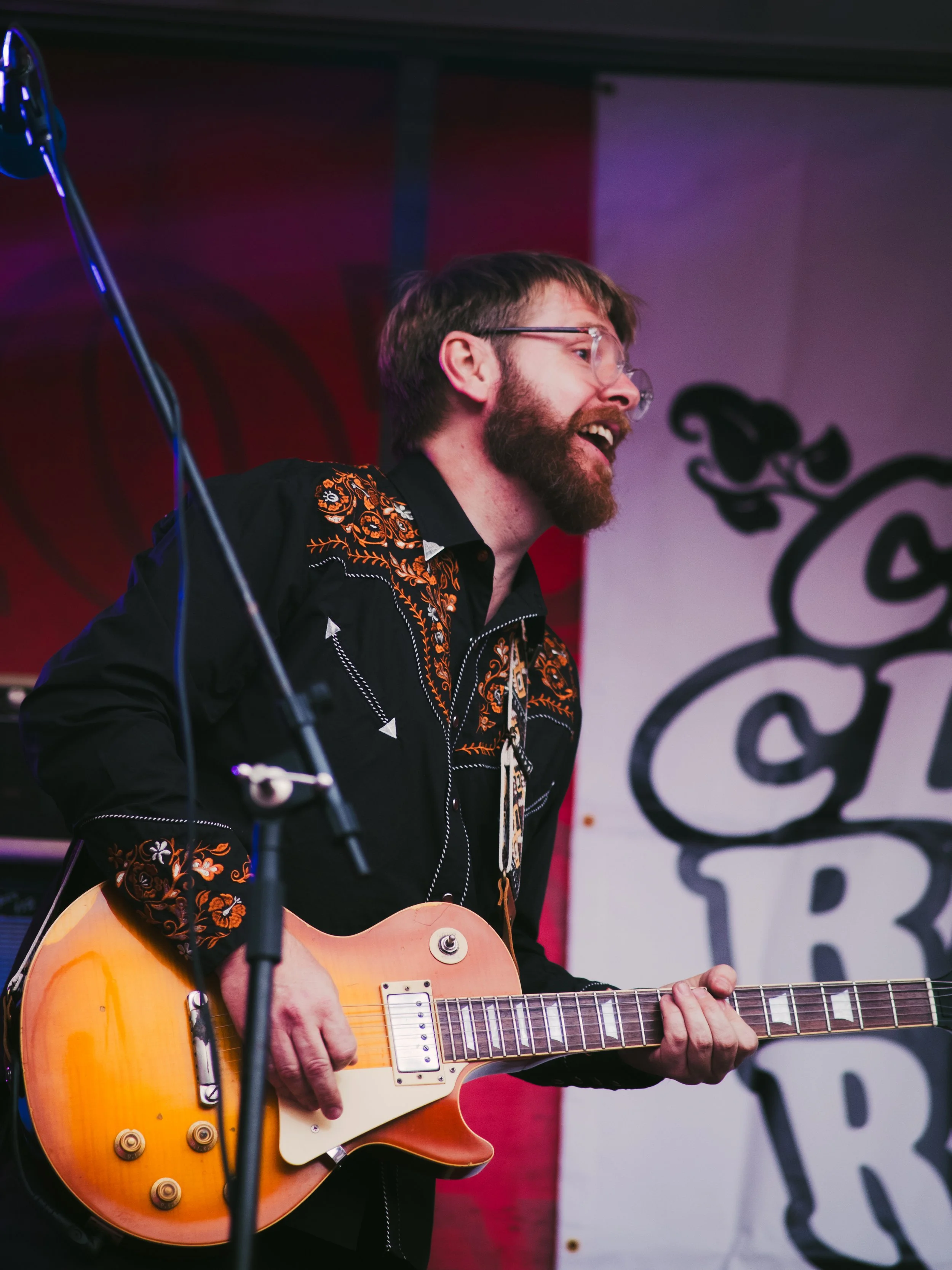 A man with glasses, a beard, and a mustache playing an electric guitar on stage during a performance, wearing a black embroidered Western shirt.
