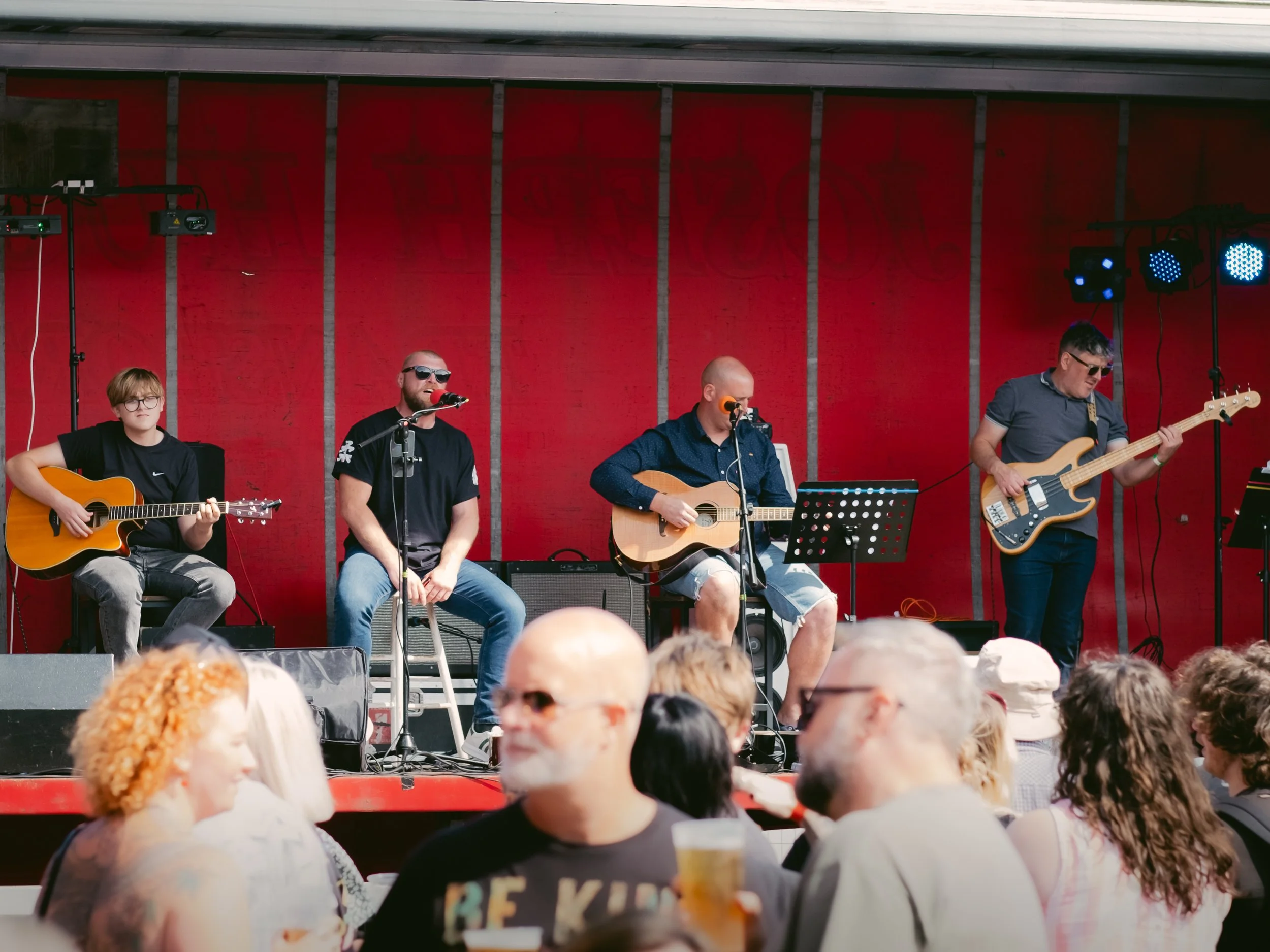 A band of four men performing on a stage with red backdrop, playing guitars and singing into microphones, in front of an audience.