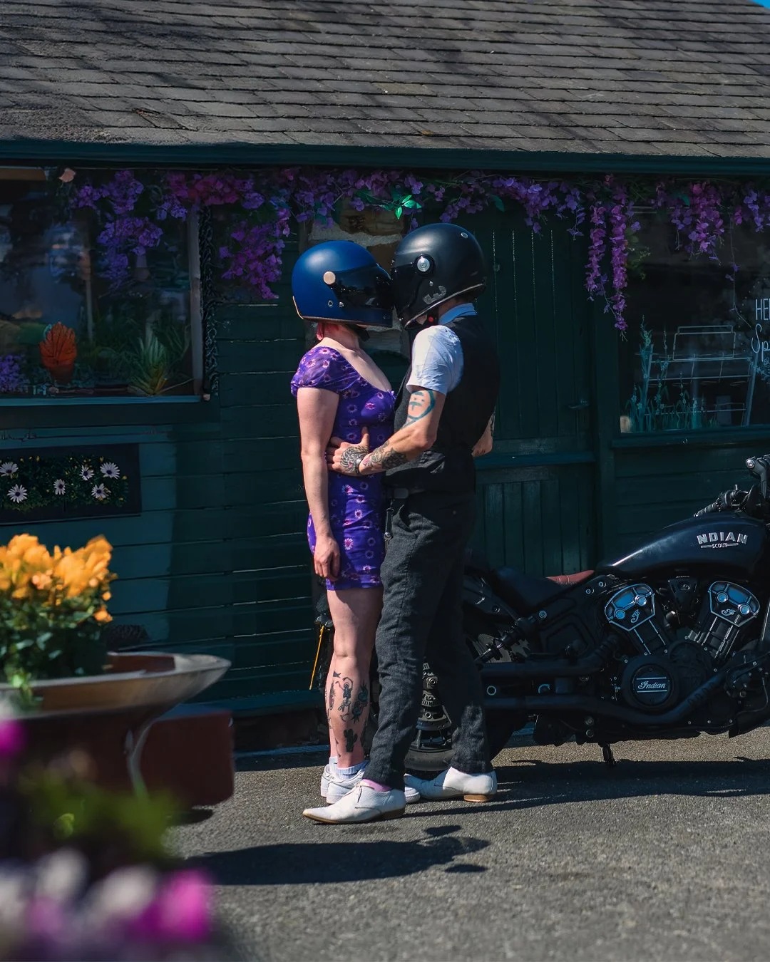 A couple wearing motorcycle helmets standing close together, about to kiss, with a black motorcycle nearby and a building decorated with purple flowers in the background.