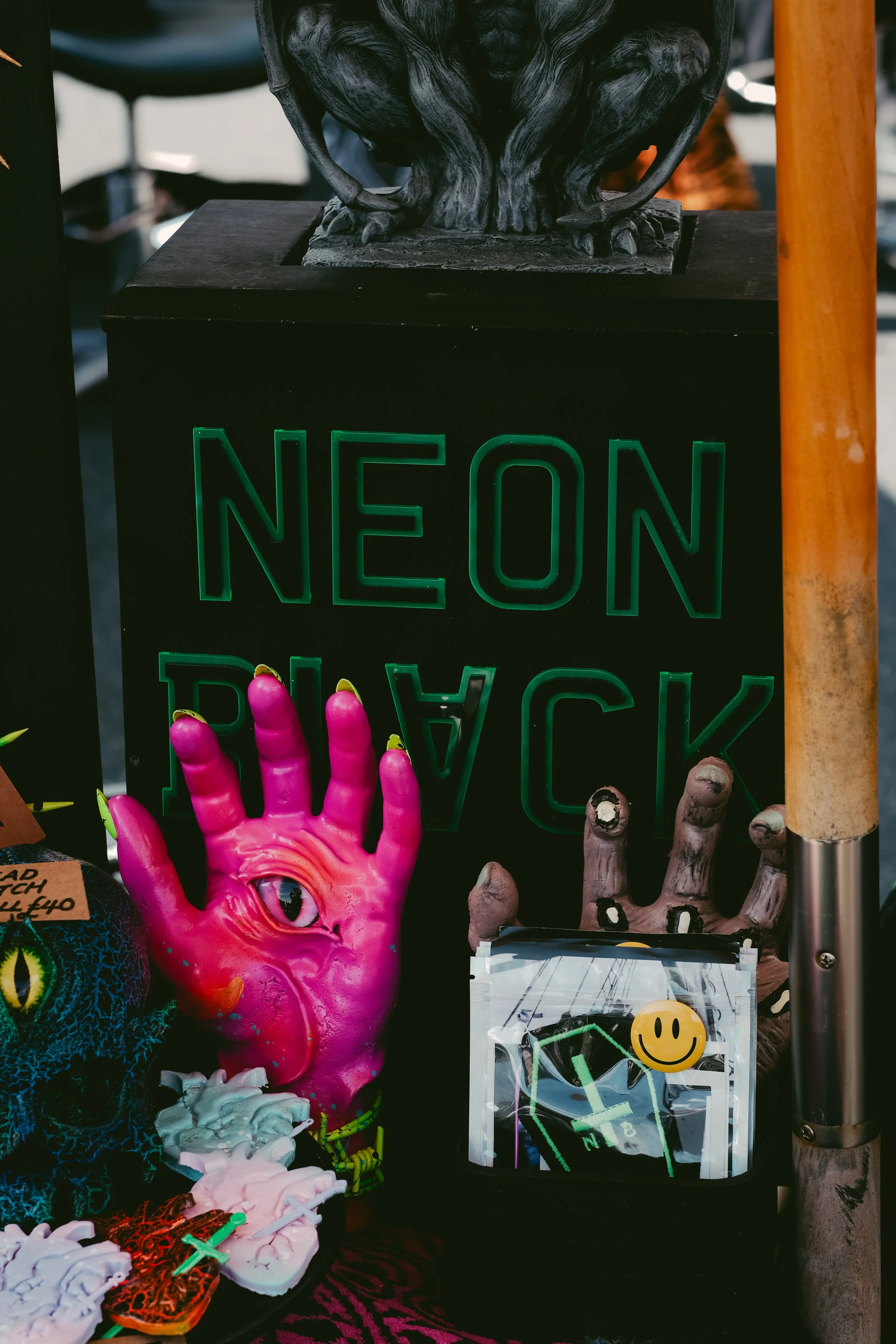 A neon sign reads 'NEON BLACK' in green letters. In front of it are artistic items, including a pink hand sculpture with an eye in the palm, a smaller hand sculpture, and various small objects including a yellow smiley face sticker.