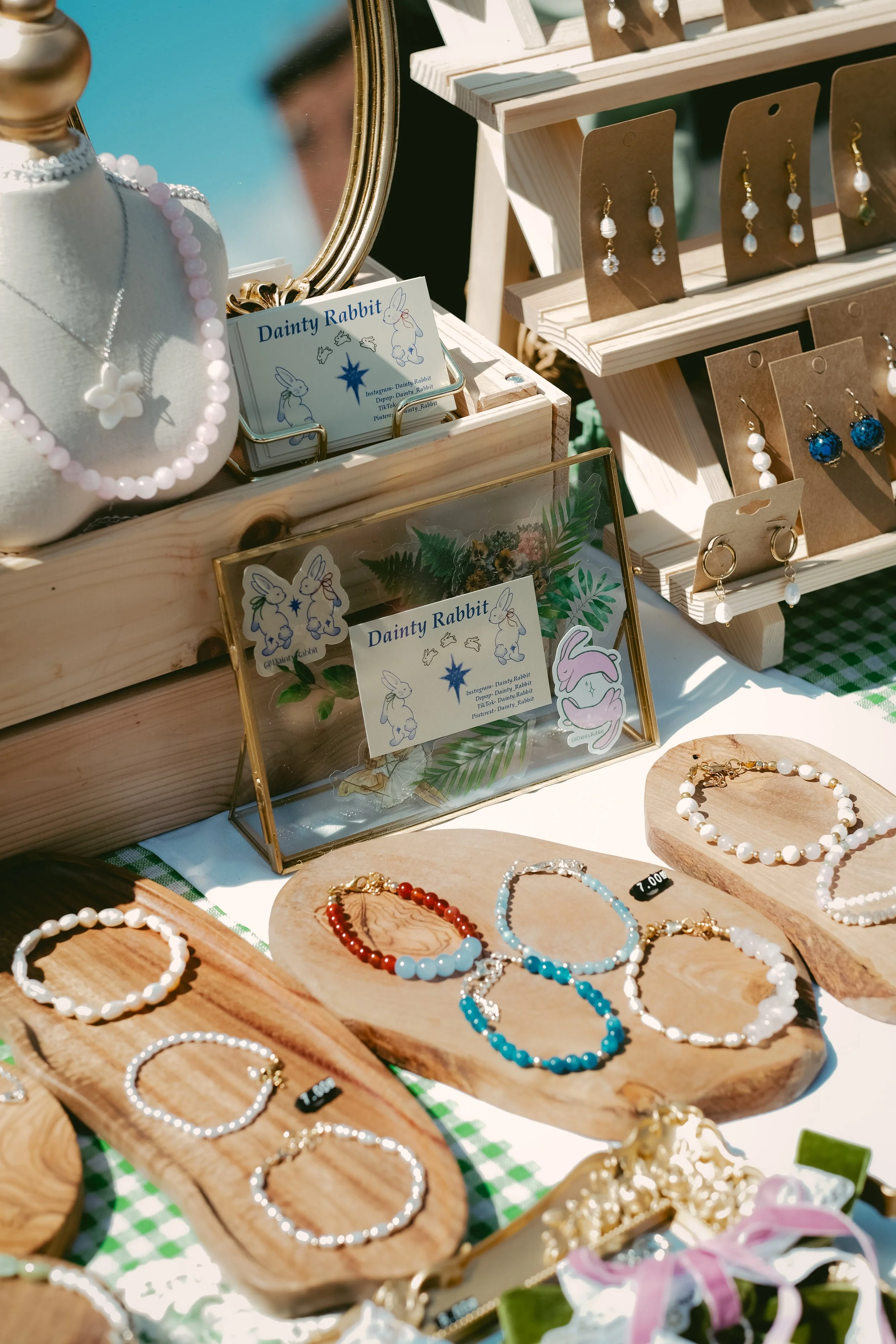 Display of jewelry including earrings, bracelets, and necklaces on wooden trays and stands at an outdoor market stall, with a hand mirror and decorative signs featuring rabbits.