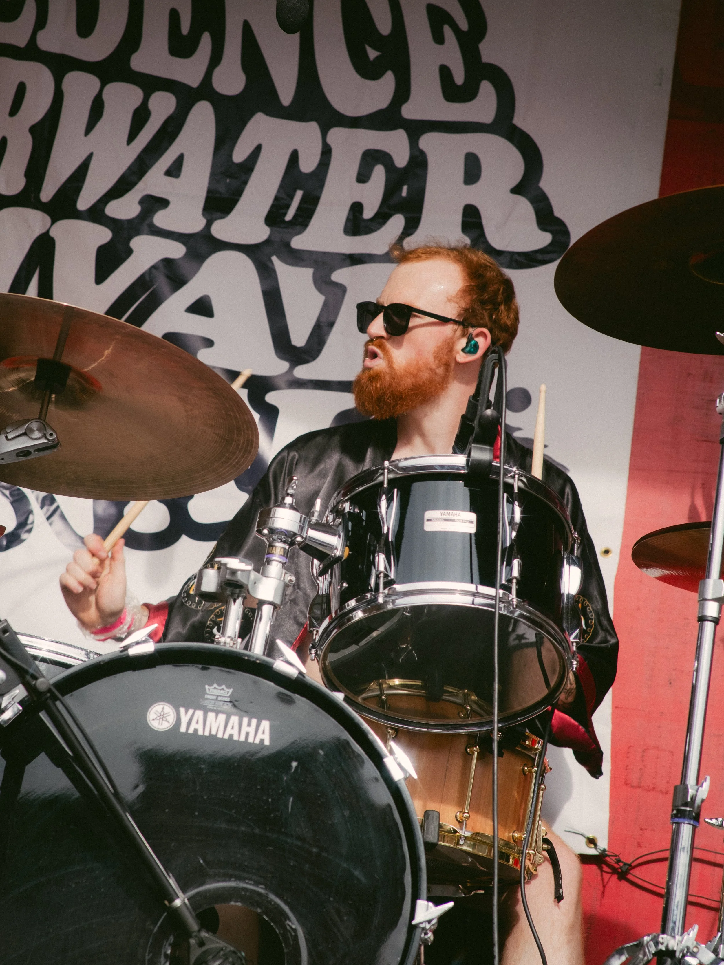 A man with red hair and beard wearing sunglasses and a black jacket playing a Yamaha drum set on stage. There is a large text logo in the background.