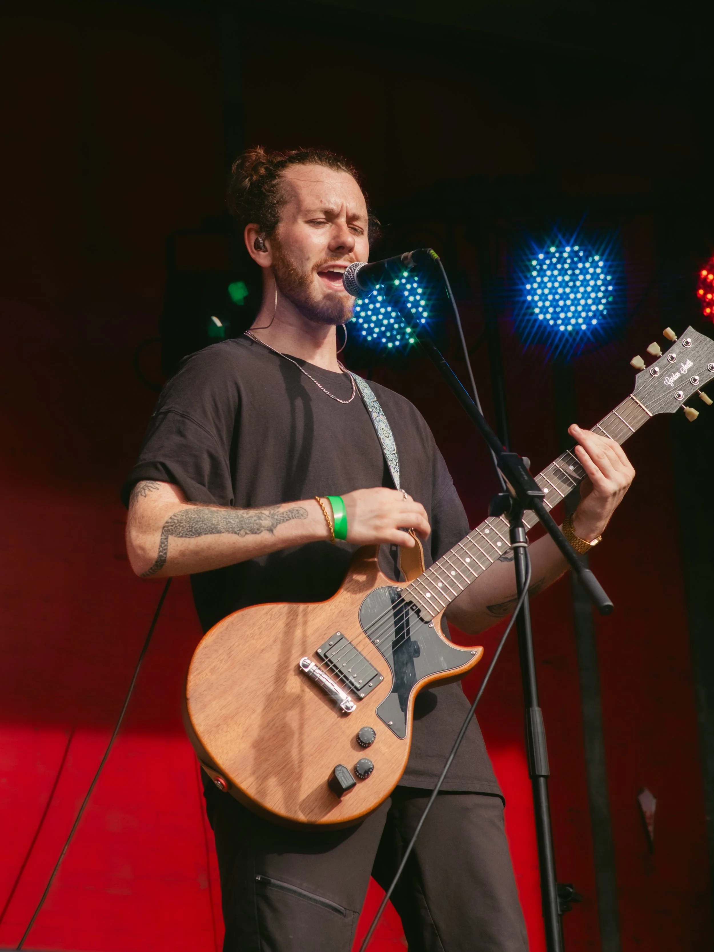 Male musician with tattoos singing and playing an electric guitar on stage, with colorful stage lights in the background.