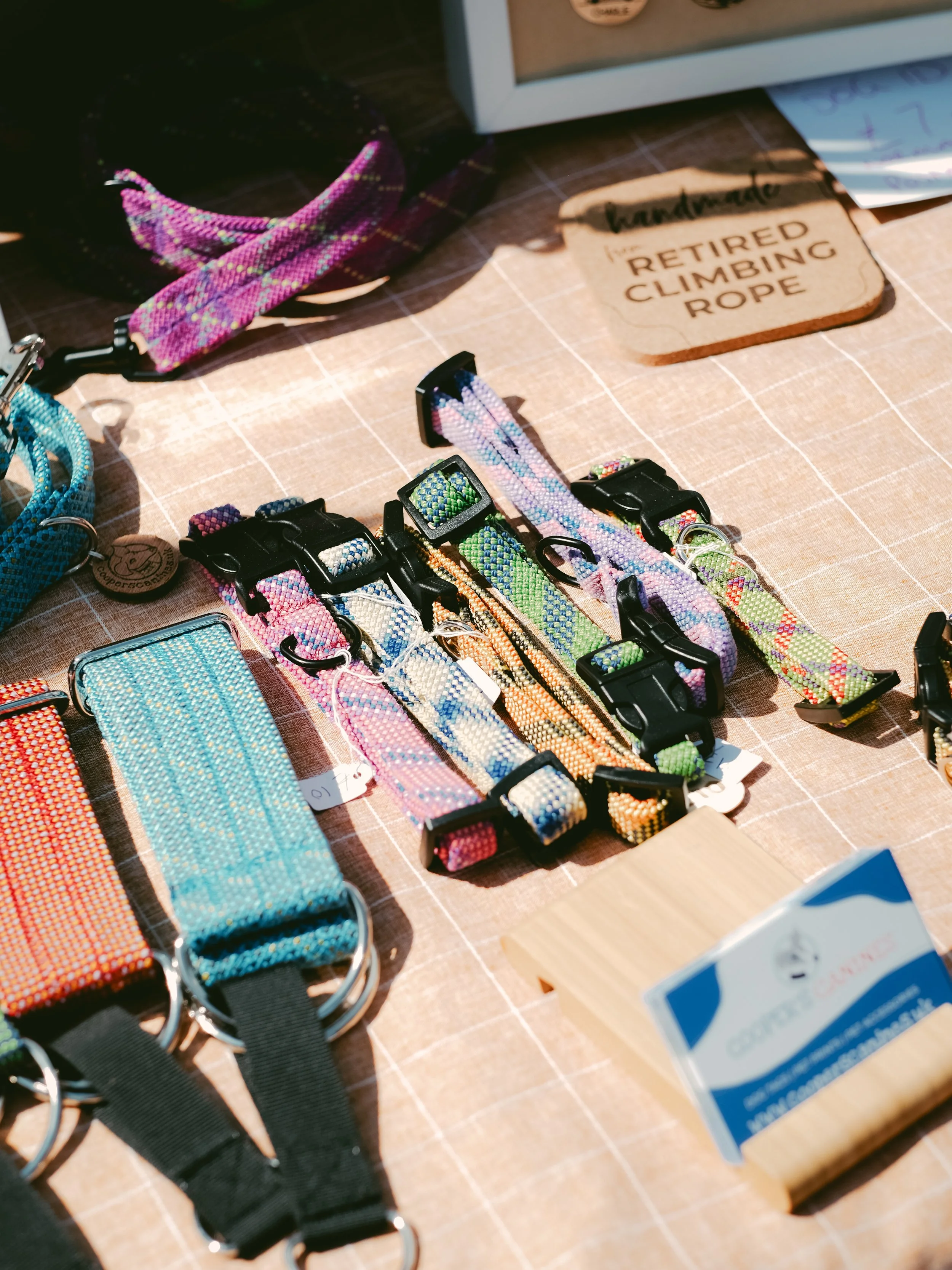 Display of colorful pet collars, leashes, and a reusable climbing rope on a table at a craft fair, with a small sign indicating they are handmade and retired climbing rope.