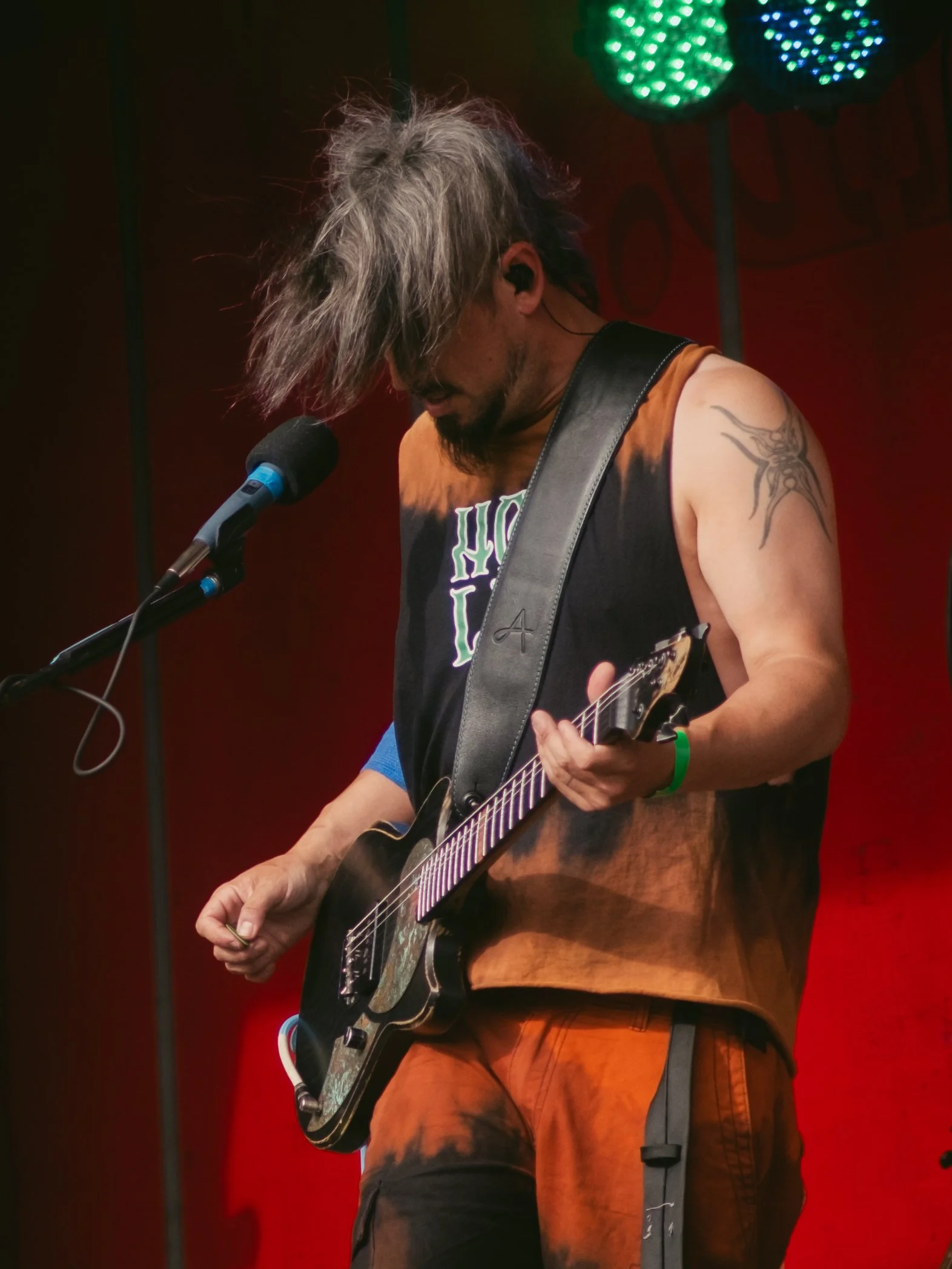 A musician performing on stage with a black electric guitar, messy hair covering his face, wearing a tank top, orange pants, and a green wristband.