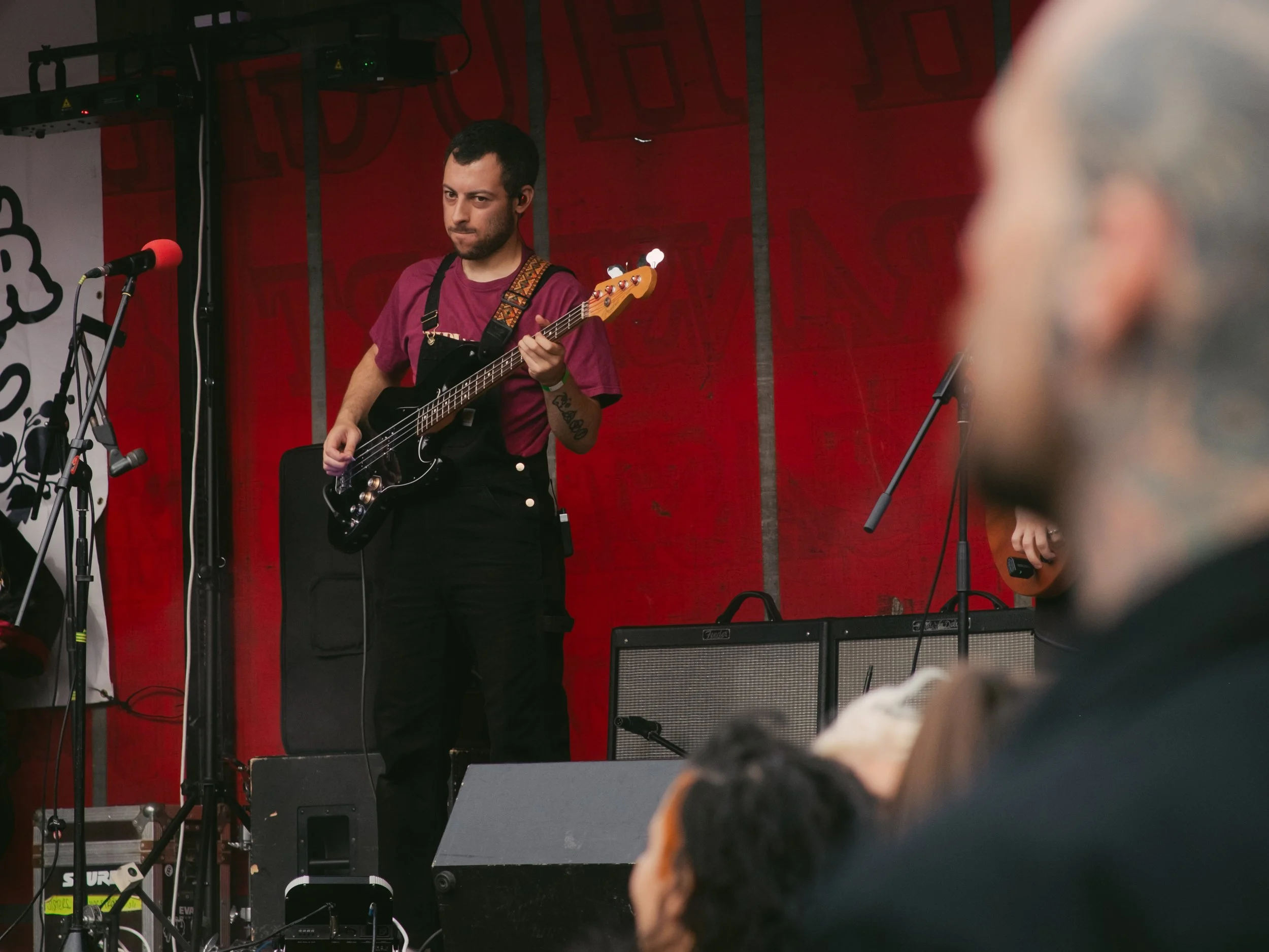 A musician playing a black electric bass guitar on stage during a live performance with a red backdrop.