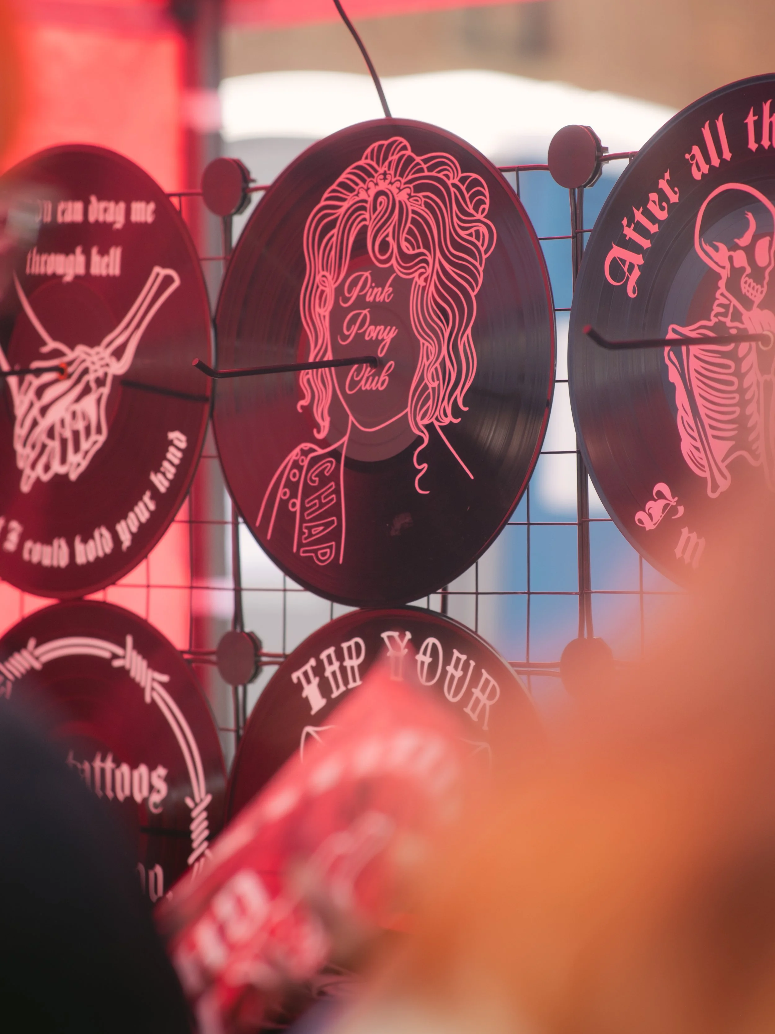 Decorative vinyl records with pink designs and text, including a central record titled "Pink Pony Club" with an illustration of a woman with curly hair, mounted on a wire display.