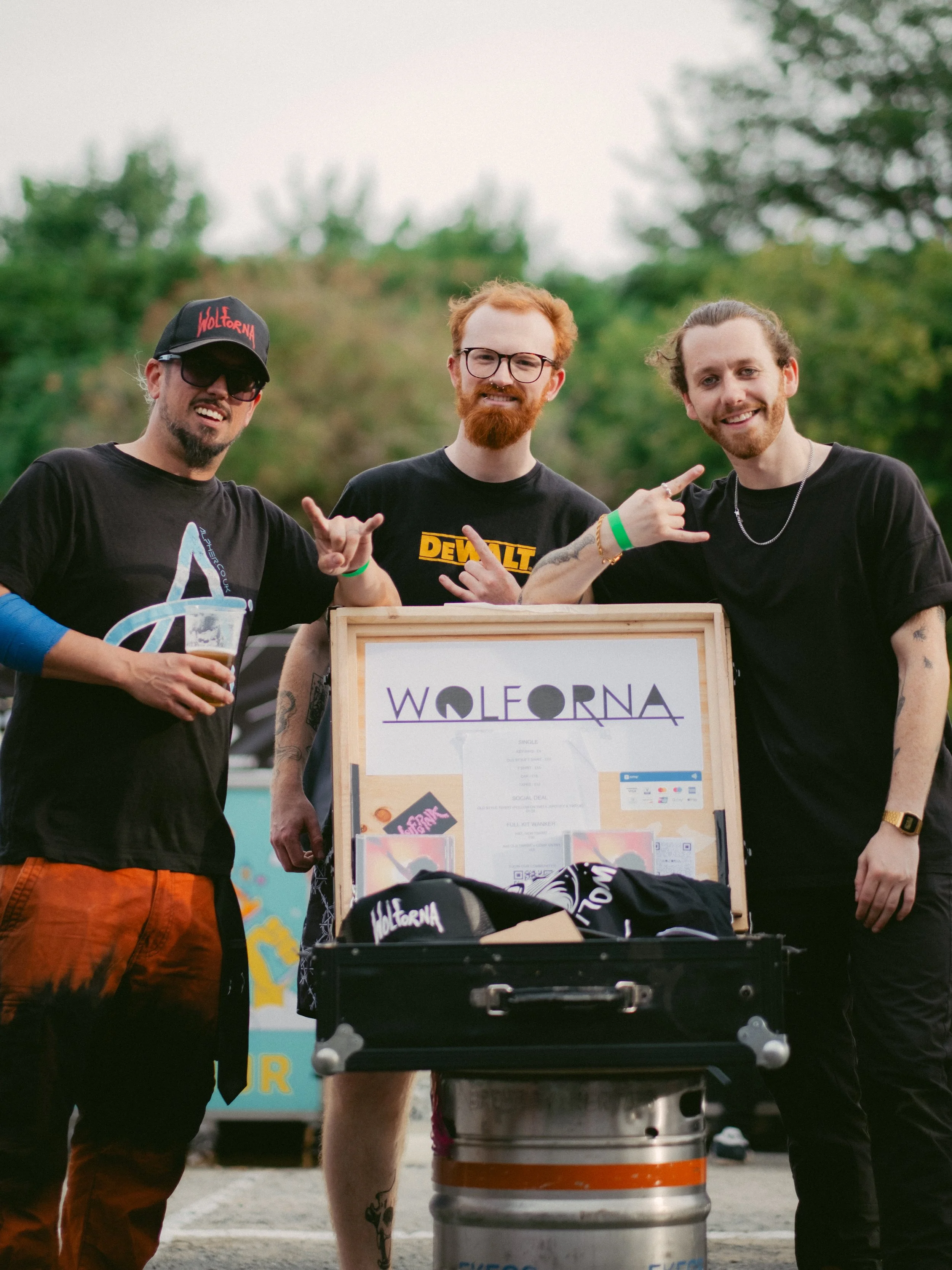 Three men standing outdoors at a festival, behind a booth with a sign titled 'WOLERNA'. One man on the left is holding a cup of beer and wearing a black cap, sunglasses, and a black T-shirt. The man in the middle has red hair, glasses, a red beard, a
