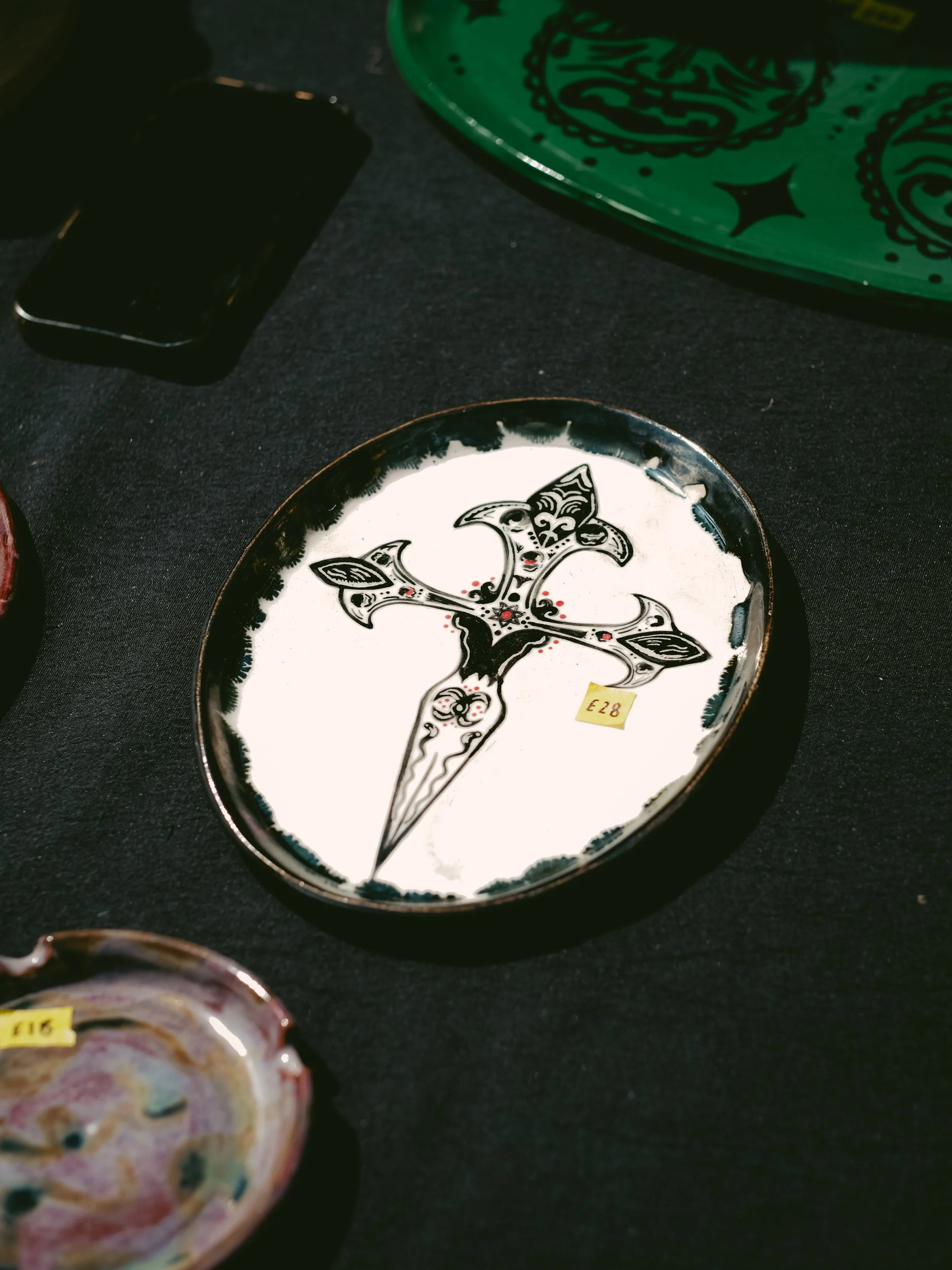 Decorative ceramic dish with a stylized dagger and floral design in black and white, priced at £28, placed on a black surface.