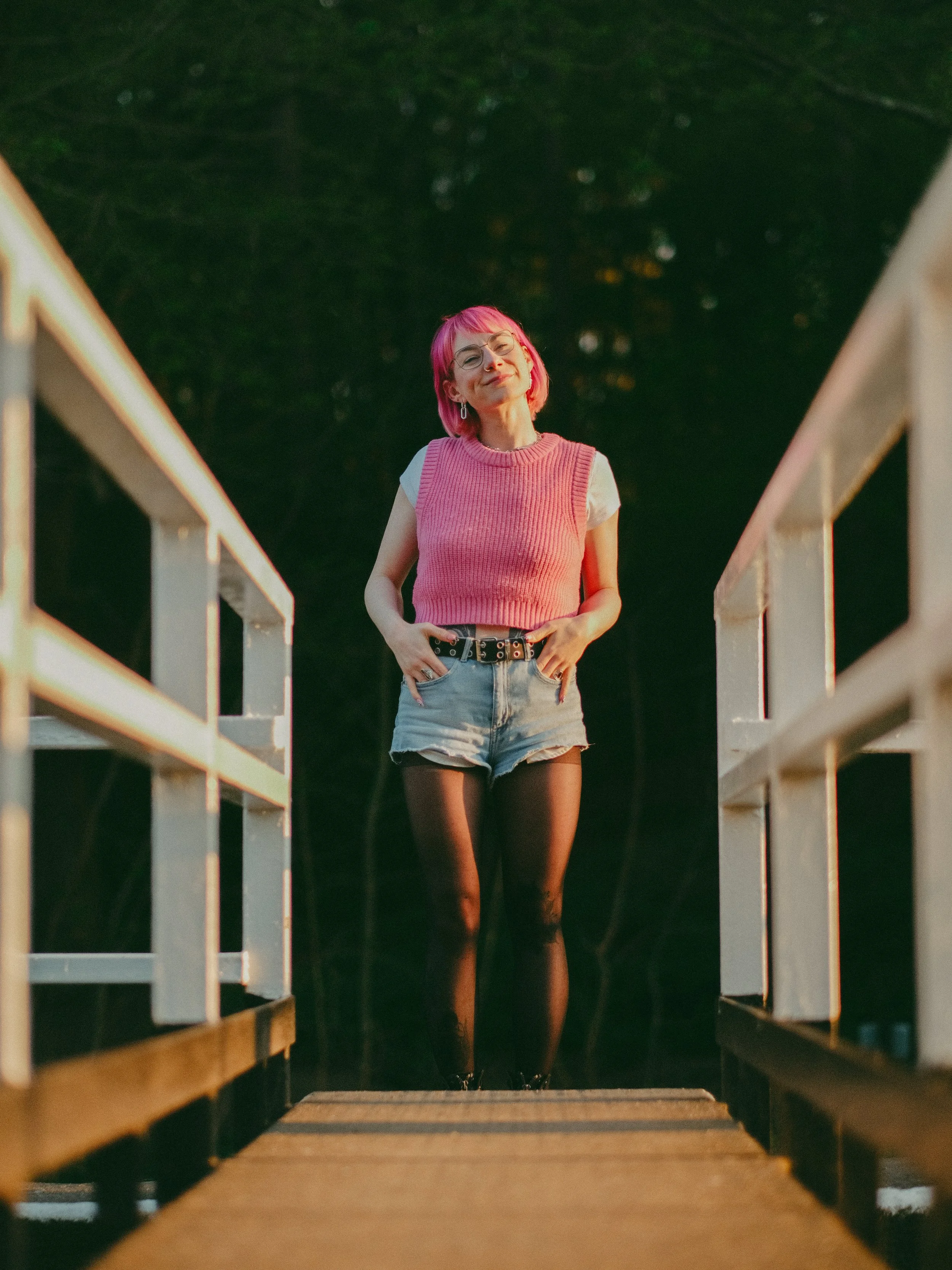 A young woman with pink hair, wearing a pink sleeveless sweater, white t-shirt, denim shorts, black tights, and black shoes, standing on a wooden bridge at sunset, smiling with her hands in her pockets.