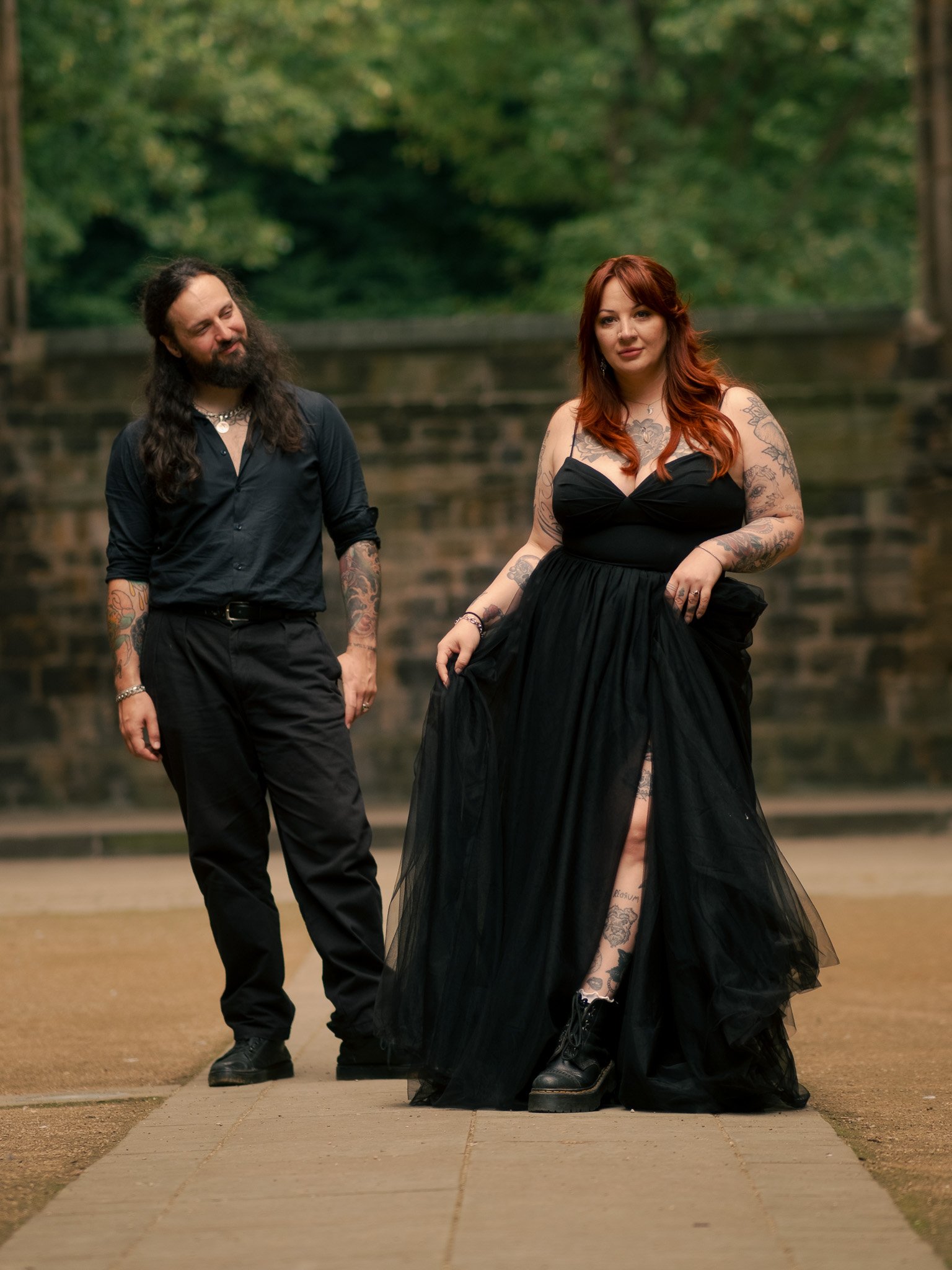 A woman with red hair and tattoos wearing a black dress and boots, walking on a path, with a man with long dark hair and tattoos in a black shirt and pants standing behind her, outdoors with trees and a stone wall in the background.