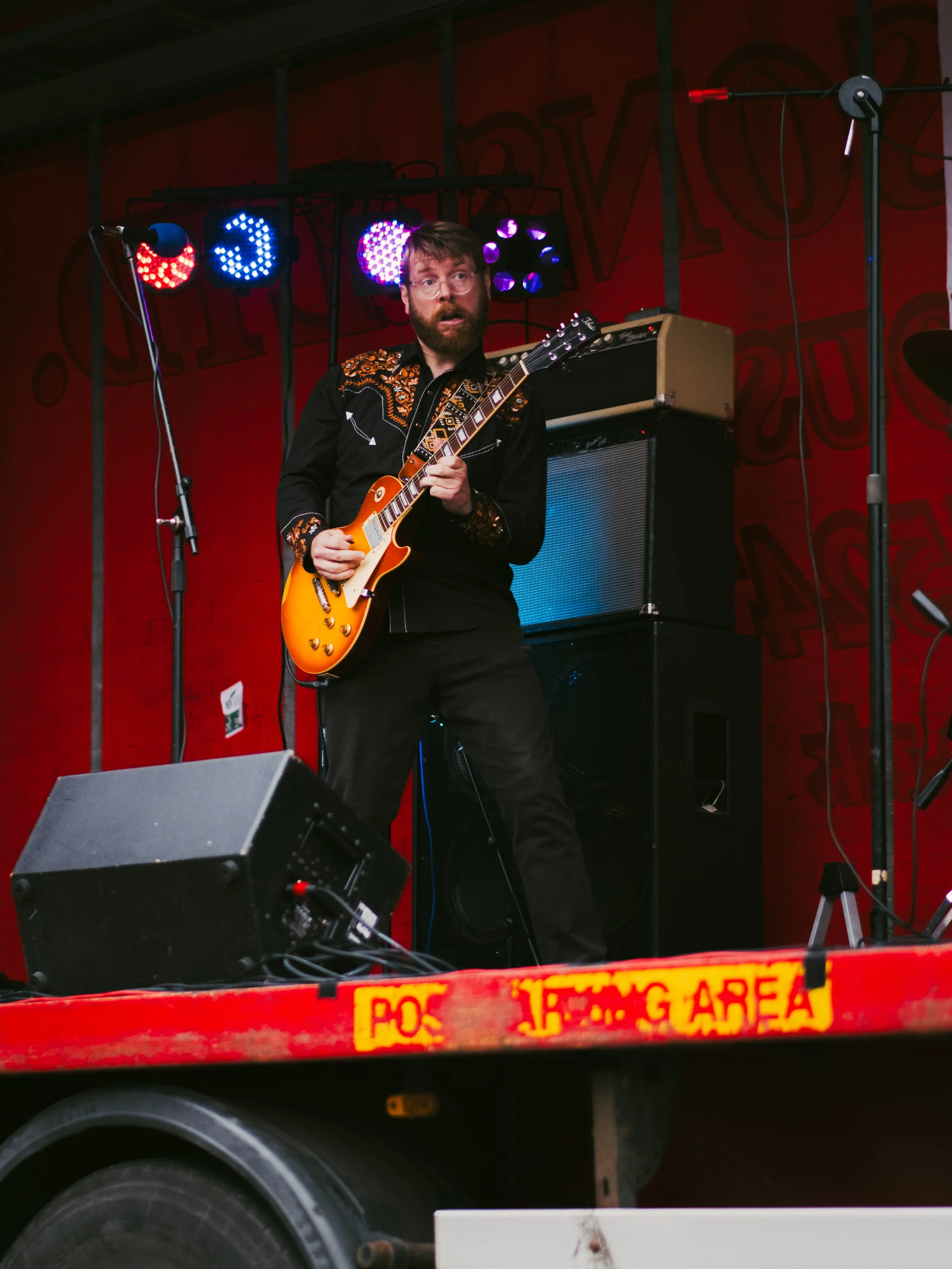 A man playing an electric guitar on a stage with red walls and colorful stage lights.