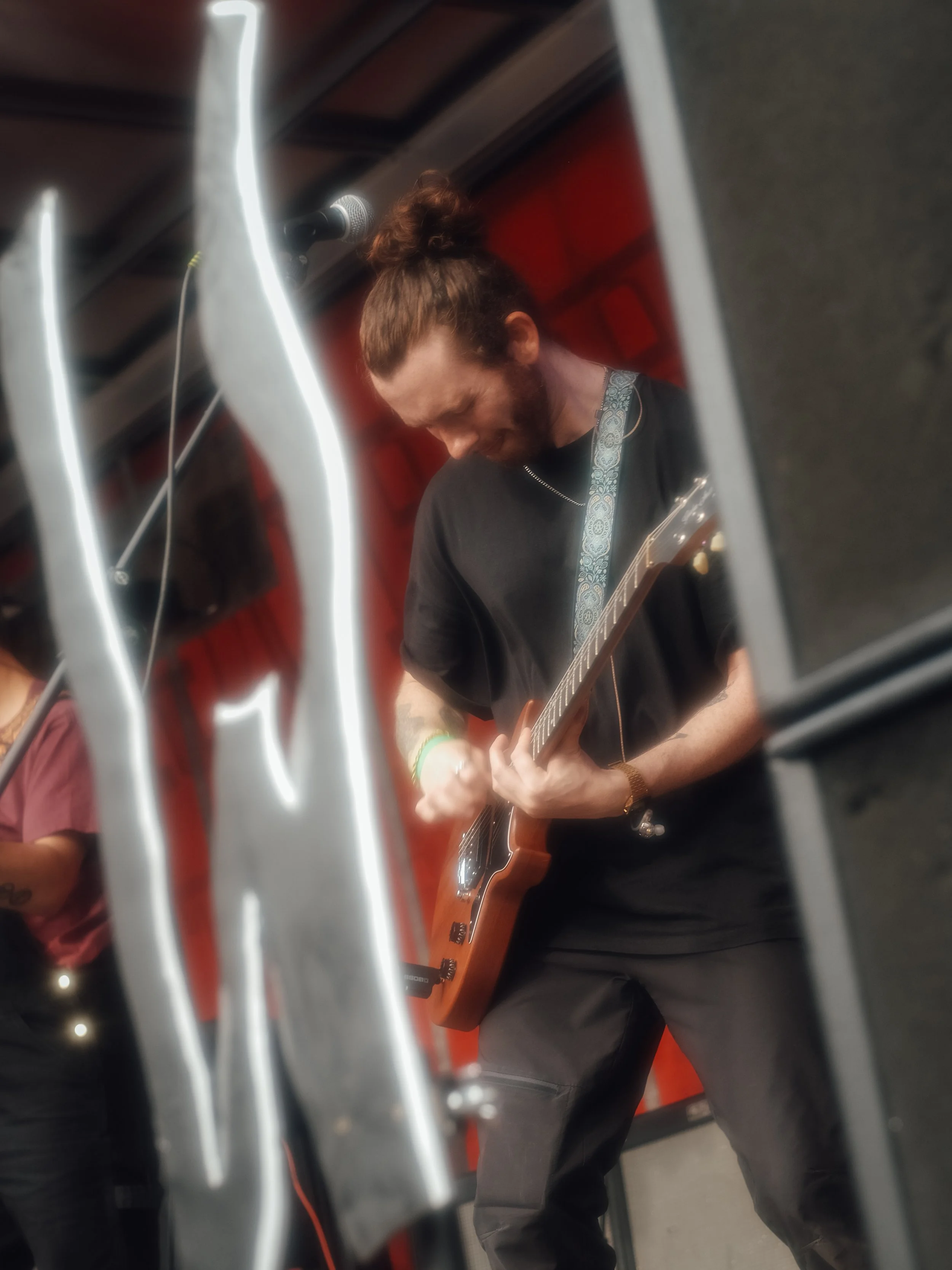 A man with a beard and long hair tied back, wearing a black T-shirt, playing an electric guitar on stage with a red background.
