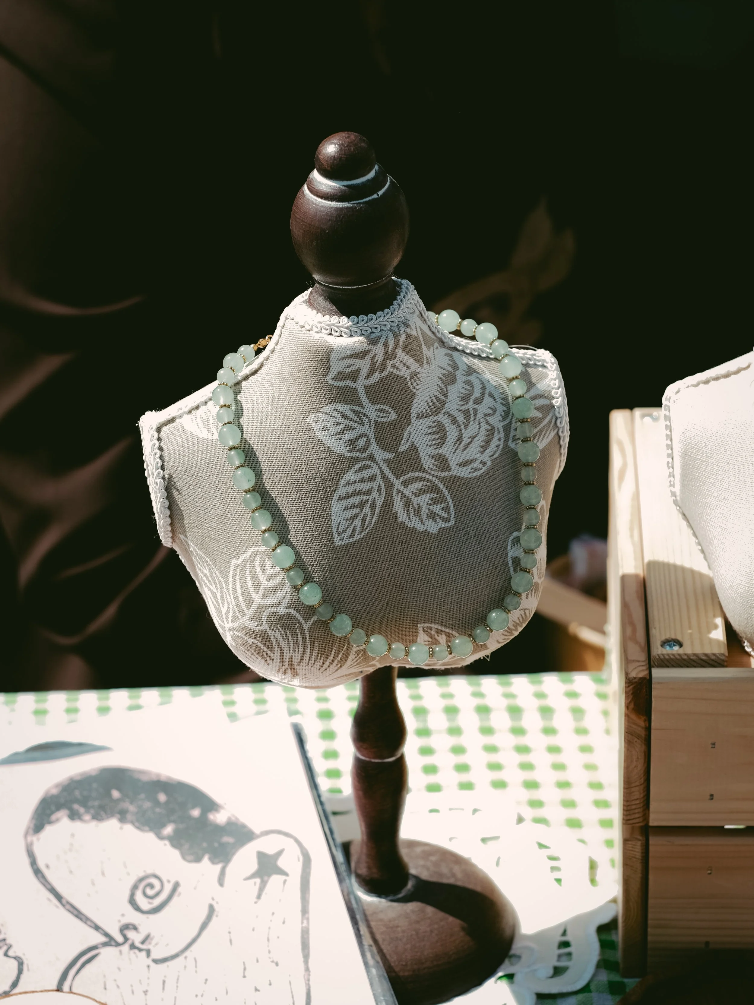 Jewelry displayed on a fabric-covered bust, with a necklace featuring round beads, set on a wooden stand with a dark background.