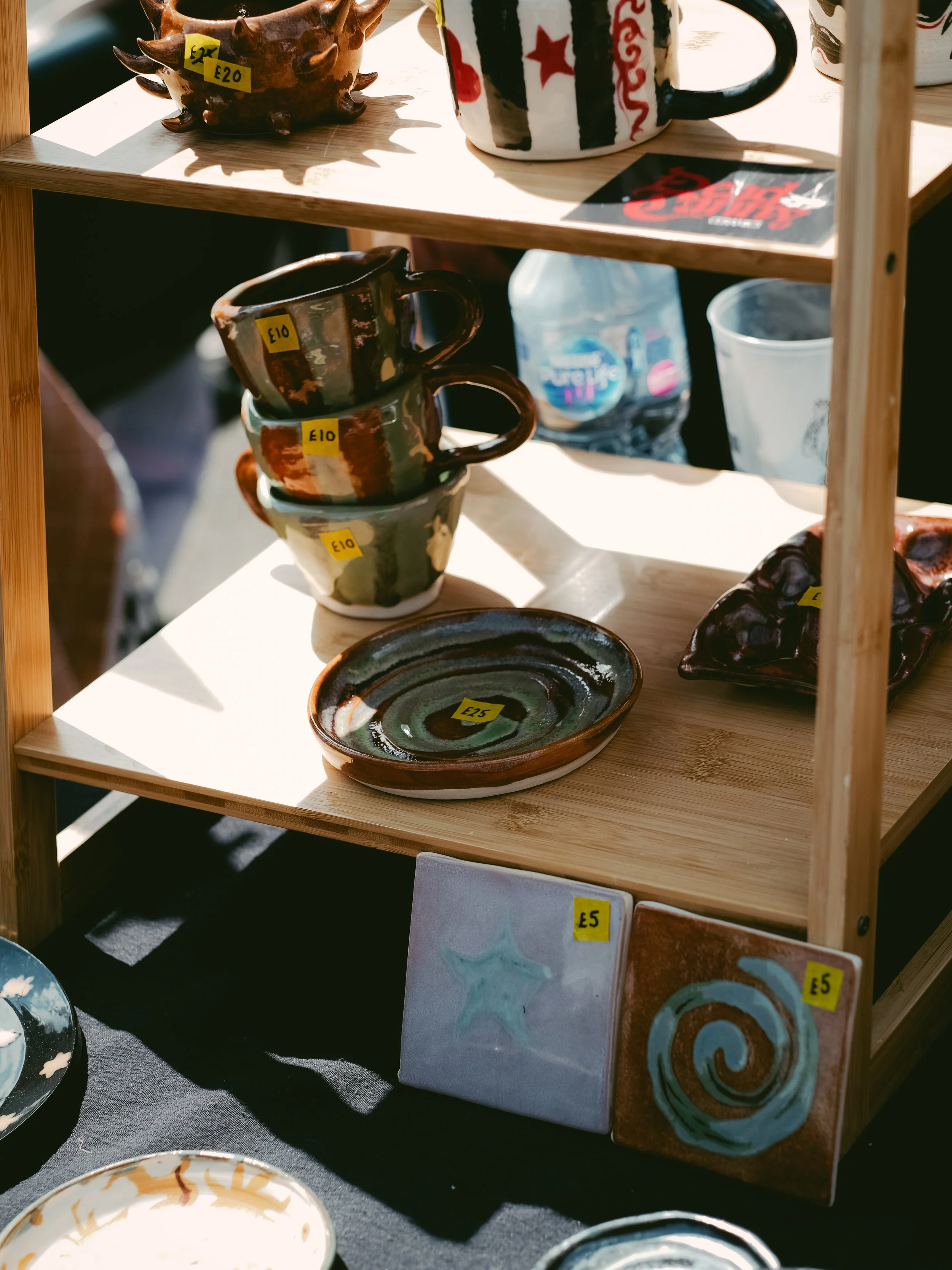 Handcrafted ceramic mugs, bowls, and tiles with prices displayed on yellow stickers, arranged on a wooden display shelf for sale.
