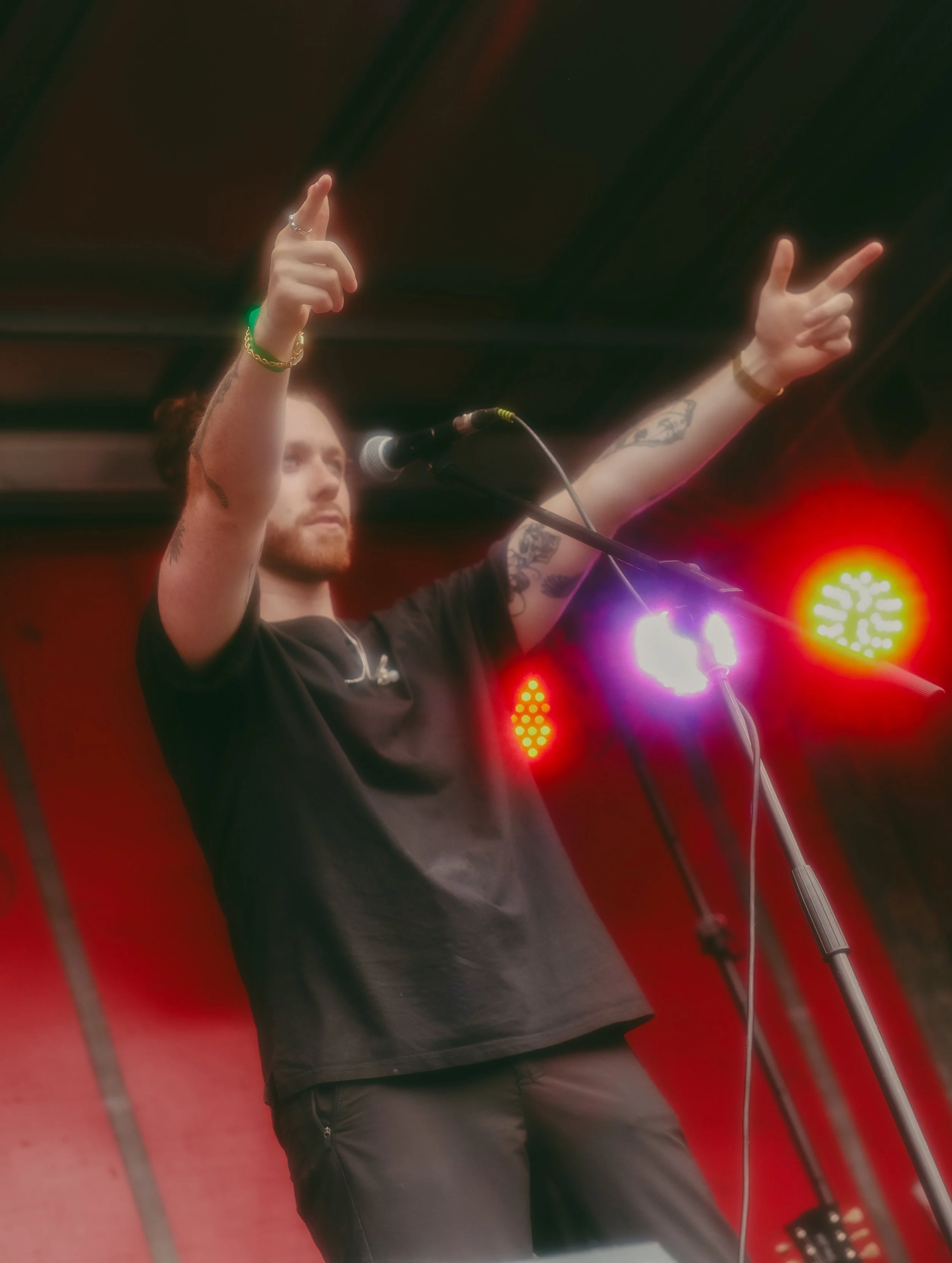 A man with tattoos on his arms, wearing a black shirt, is standing on stage with arms raised. There is a microphone in front of him, and colorful stage lights are visible in the background.
