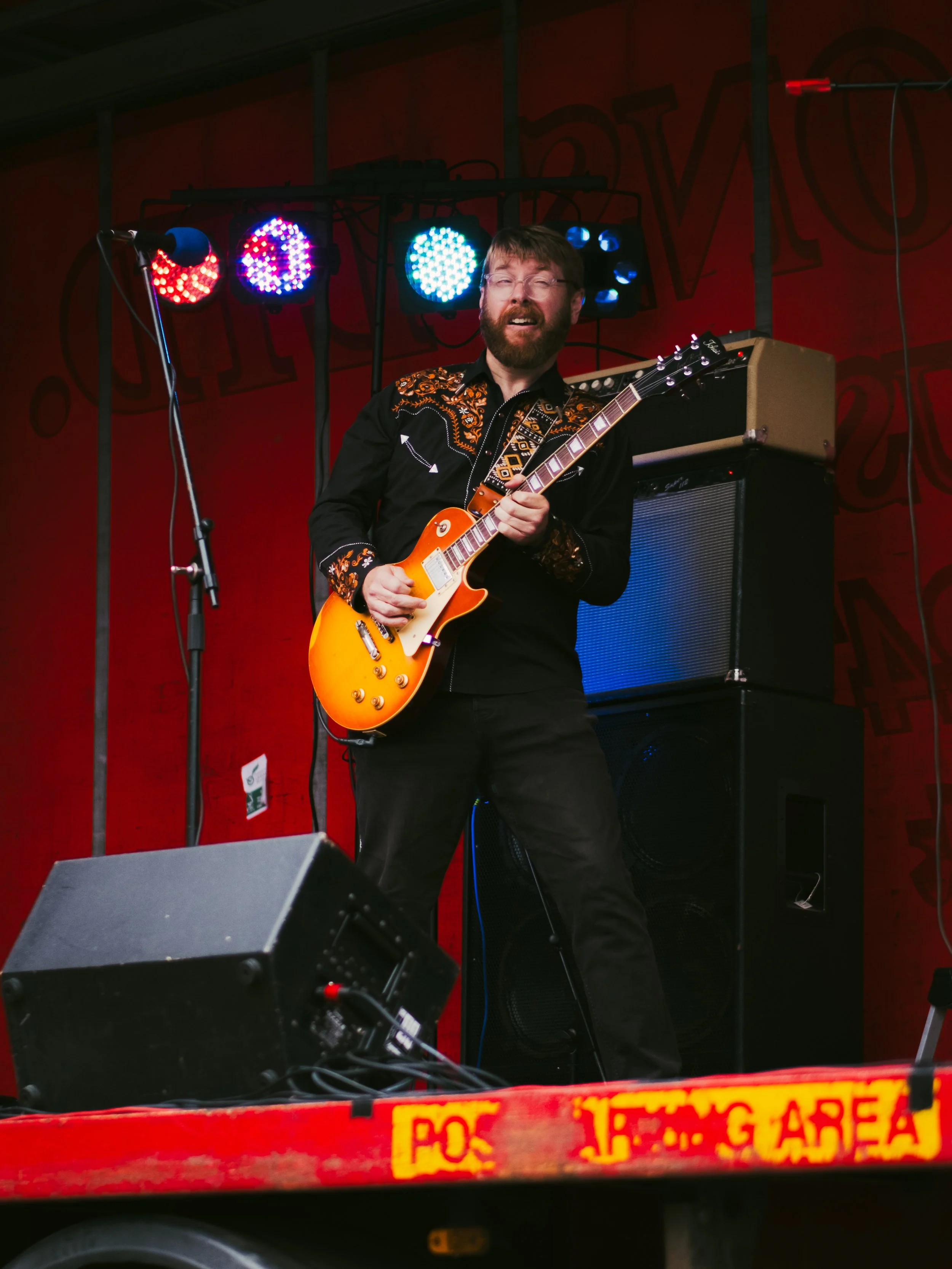 A man with glasses and a beard playing an orange electric guitar on stage with colorful lights overhead.