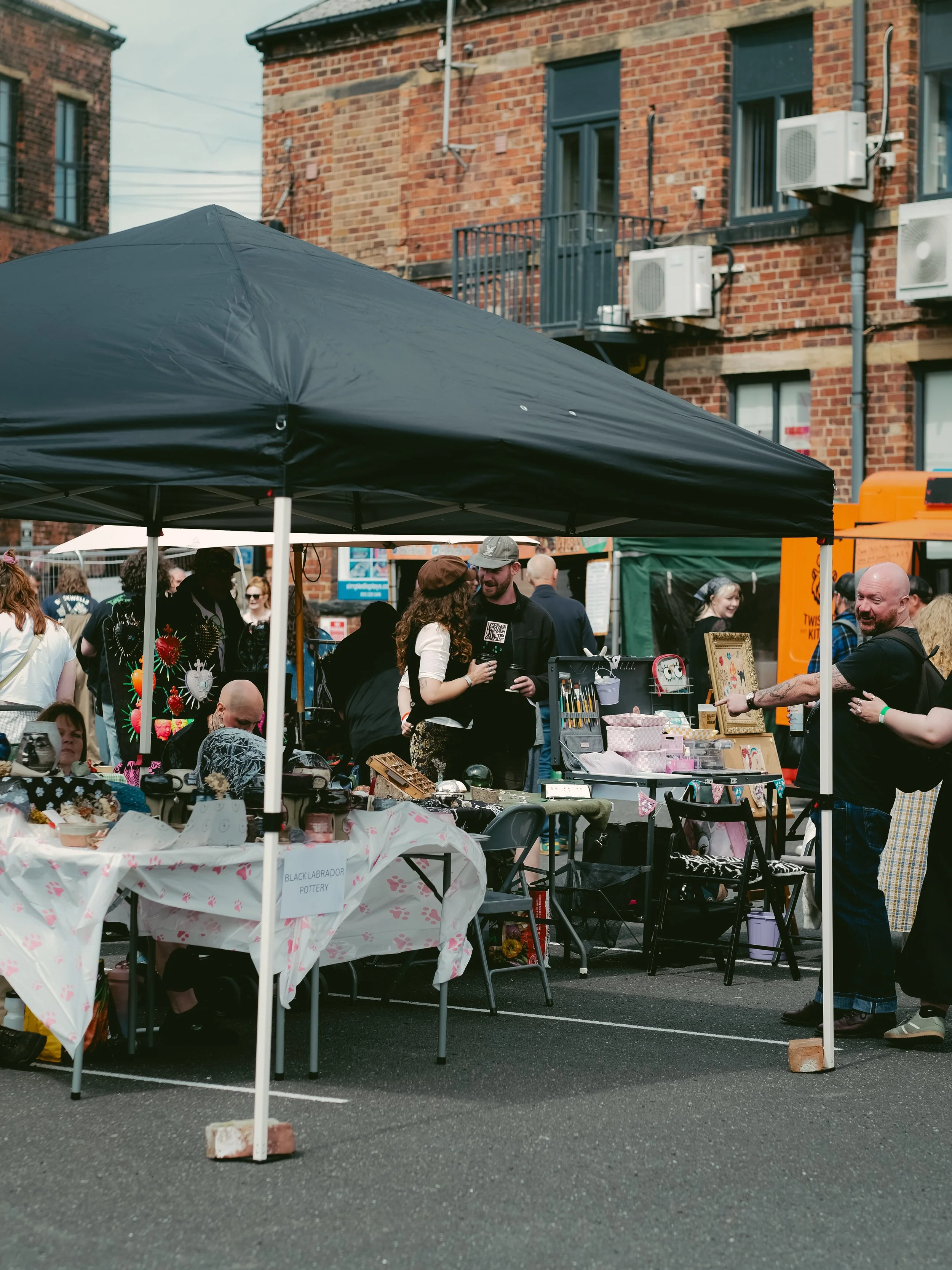 Outdoor street market with vendors displaying handmade crafts and pottery under tents, with people browsing and talking in front of brick apartment buildings.