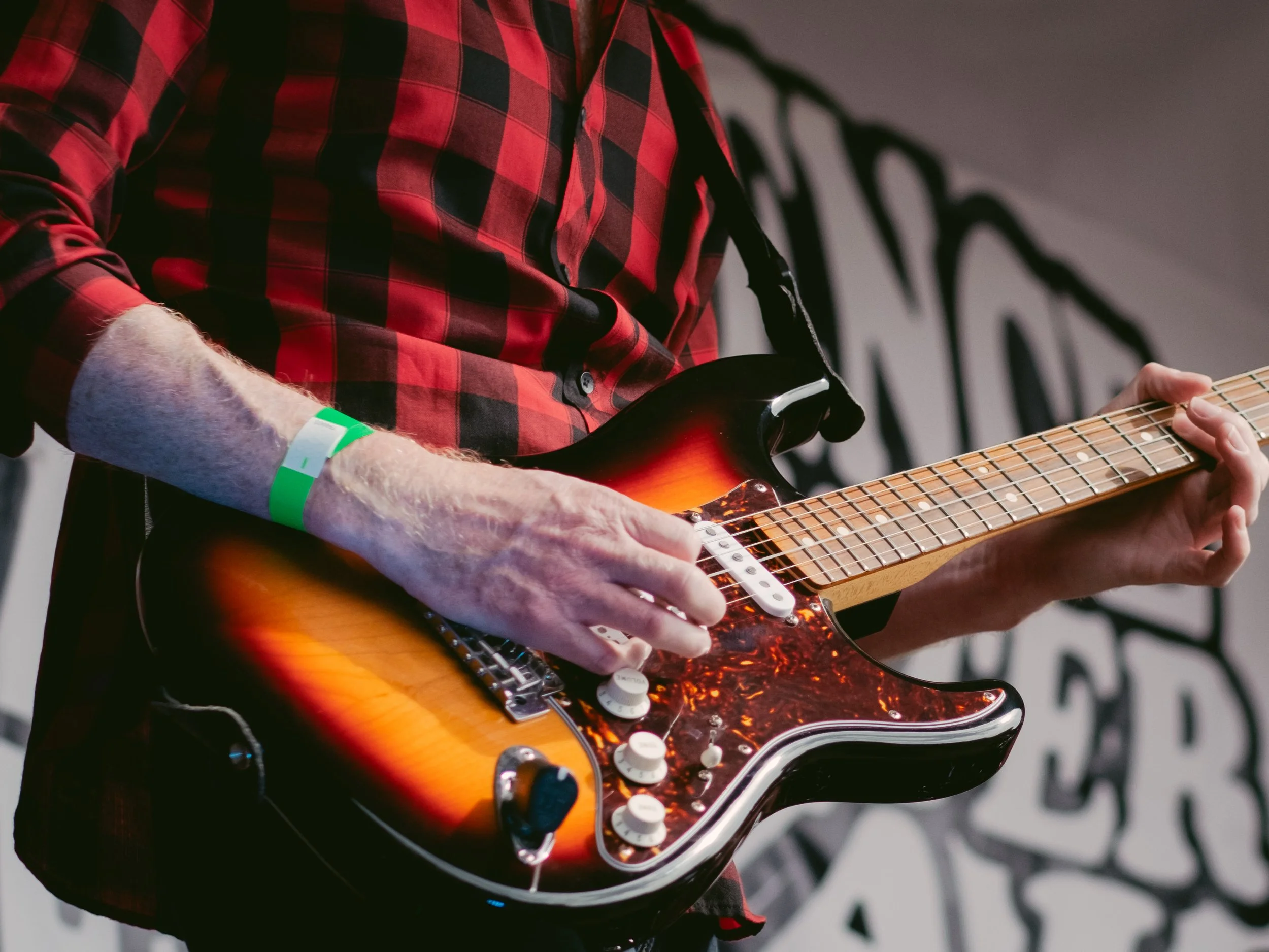 Person wearing a red and black checkered shirt playing an electric guitar with a sunburst finish and tortoise pickguard.