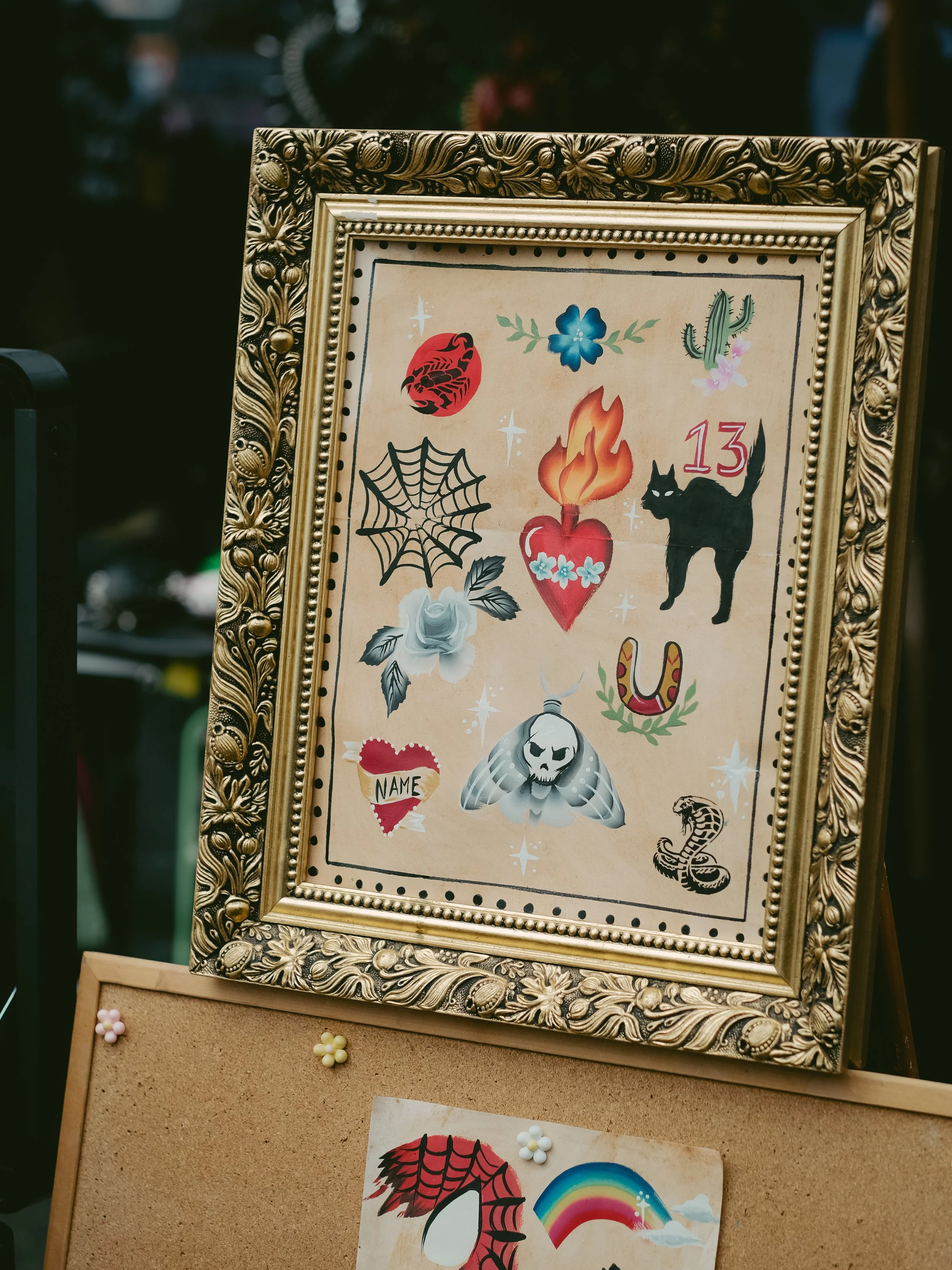 Decorative framed artwork featuring tattoos including a spider web, a black cat, a flaming heart, a skull moth, a snake, a rose, a horseshoe, and various other symbols, set against a beige background.