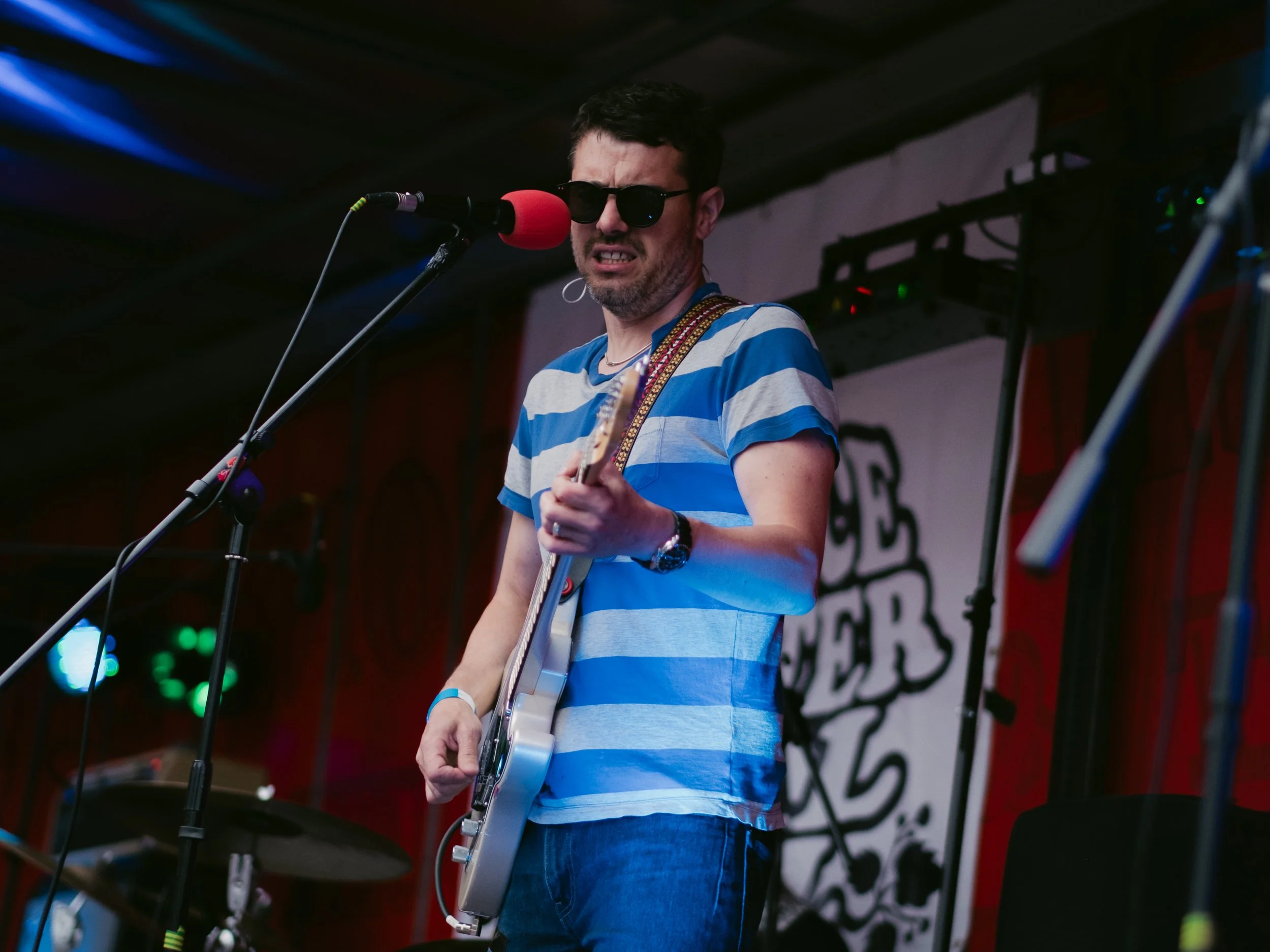 A man wearing sunglasses, a blue and white striped t-shirt, and jeans is playing an electric guitar and singing into a microphone on stage. There is a drum set in the background.