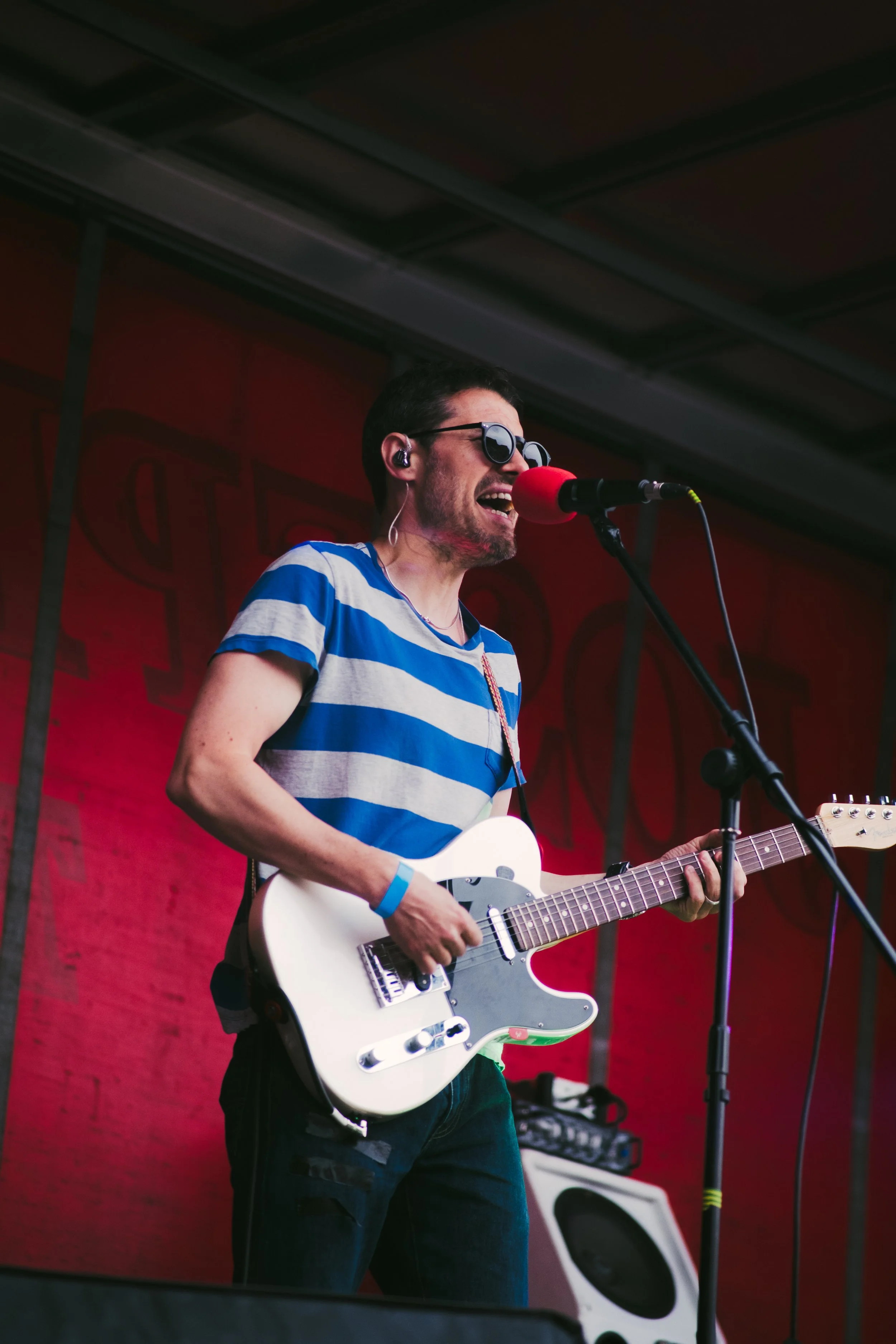 A man singing and playing electric guitar on stage, wearing sunglasses, a striped blue and gray shirt, and a blue wristband.
