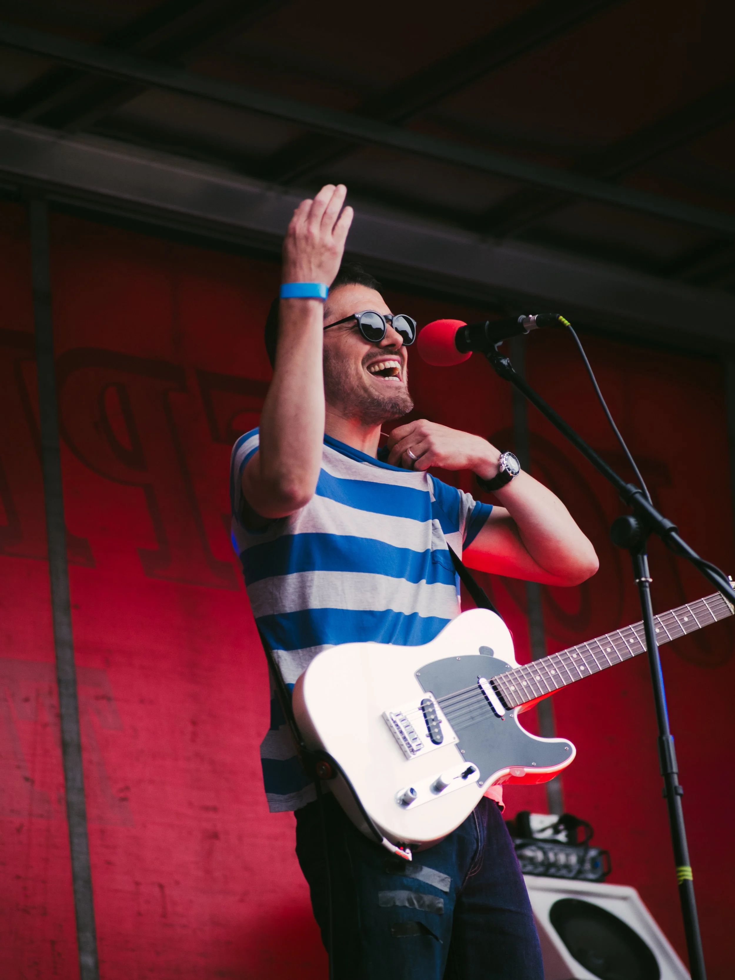 A man with sunglasses, a striped shirt, and a white electric guitar is singing and smiling at a microphone on stage.