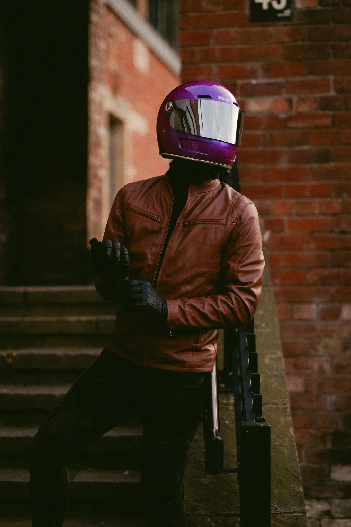Person wearing a purple motorcycle helmet and brown leather jacket standing on outdoor stairs near a brick wall.