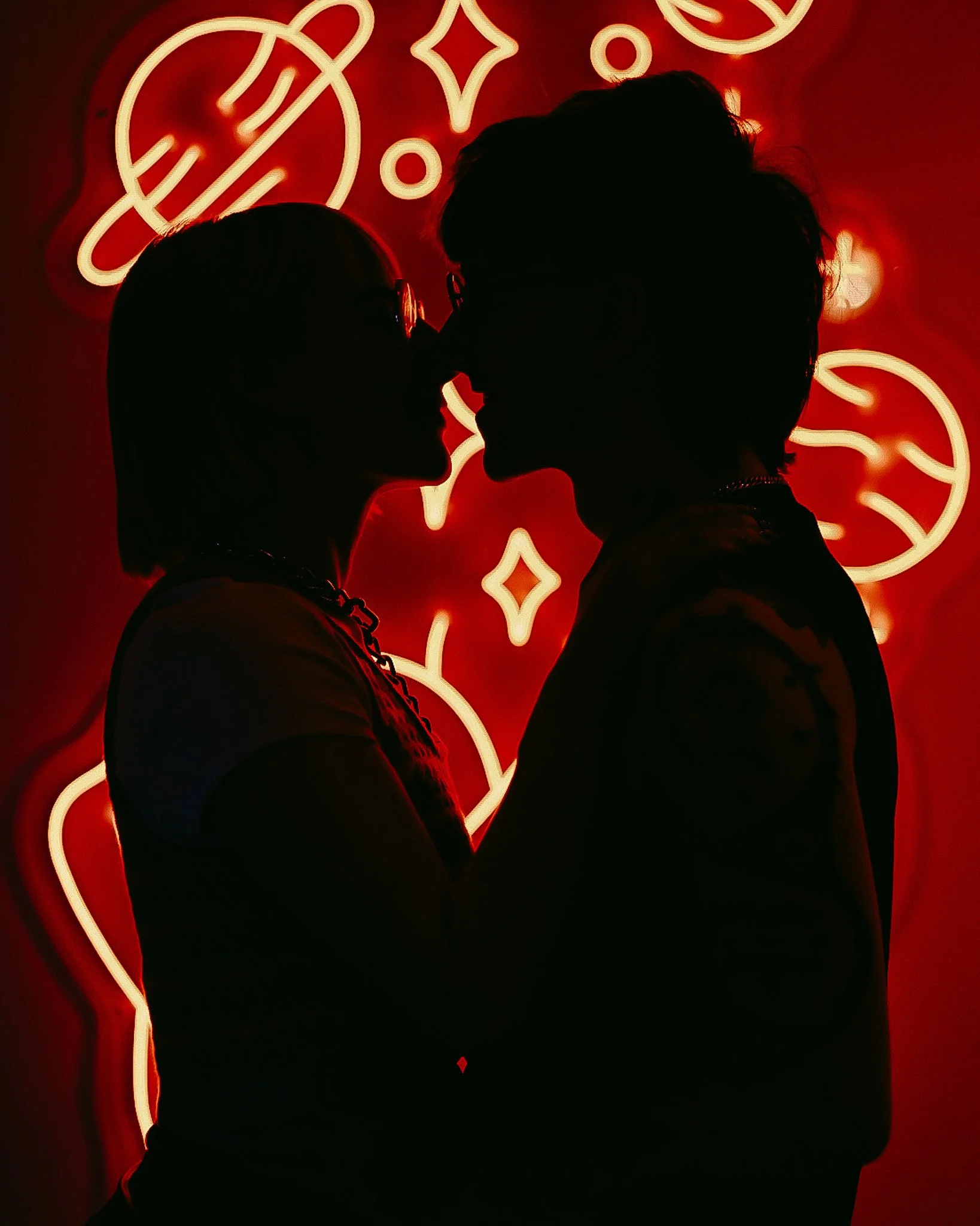 Silhouette of a man and woman facing each other close, in front of a red background with neon signs and shapes.