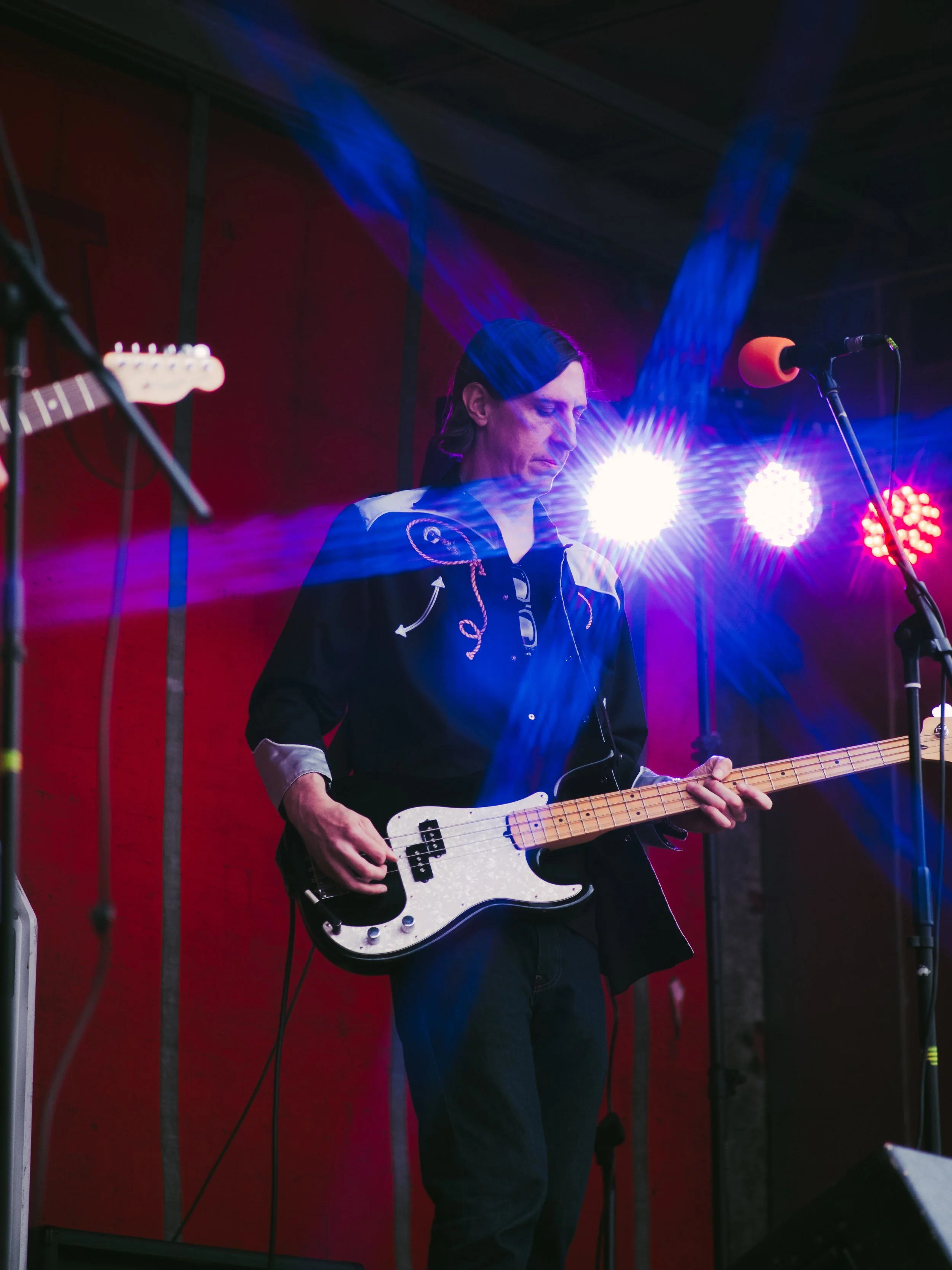 A male musician playing an electric bass guitar on stage, with colorful stage lights shining behind him.
