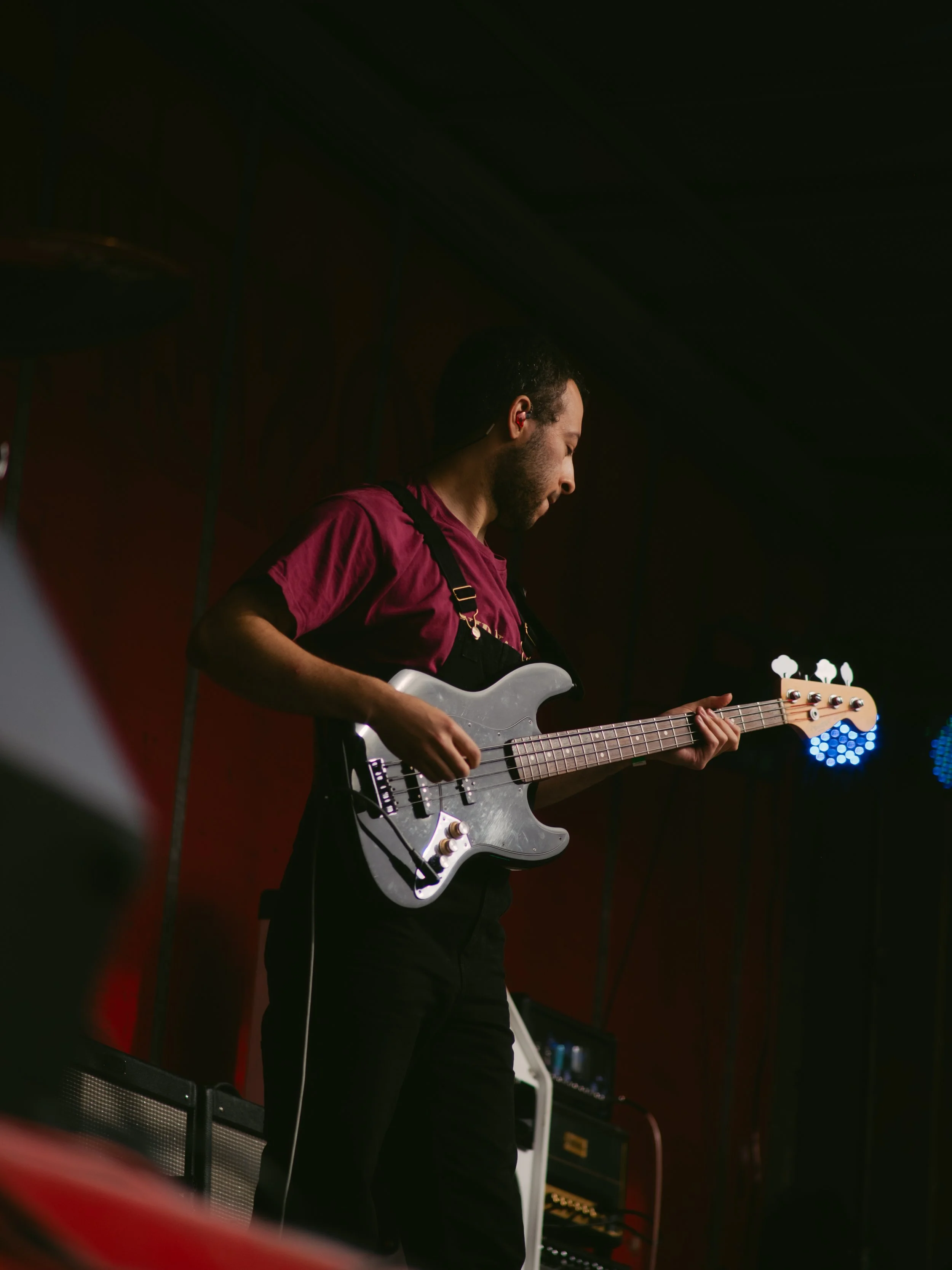 Musician playing an electric bass guitar on stage, wearing a maroon shirt and black overalls, with stage equipment visible in the background.