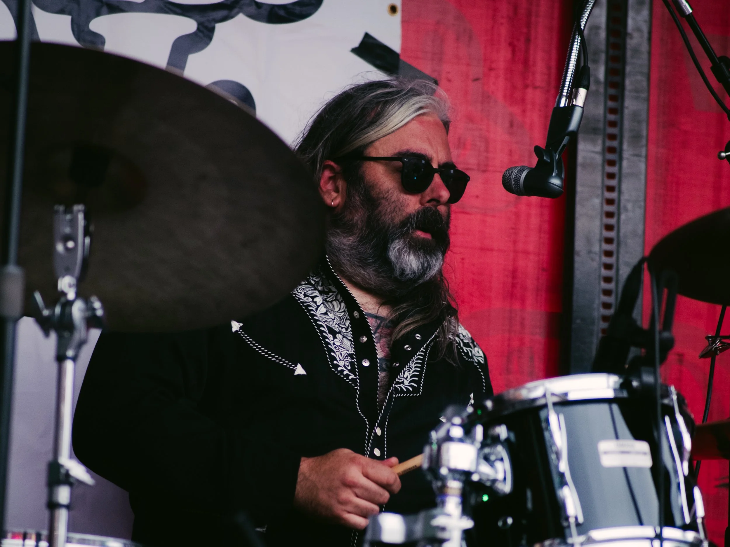 A man with long hair and a beard, wearing sunglasses, playing drums on stage with a red backdrop.
