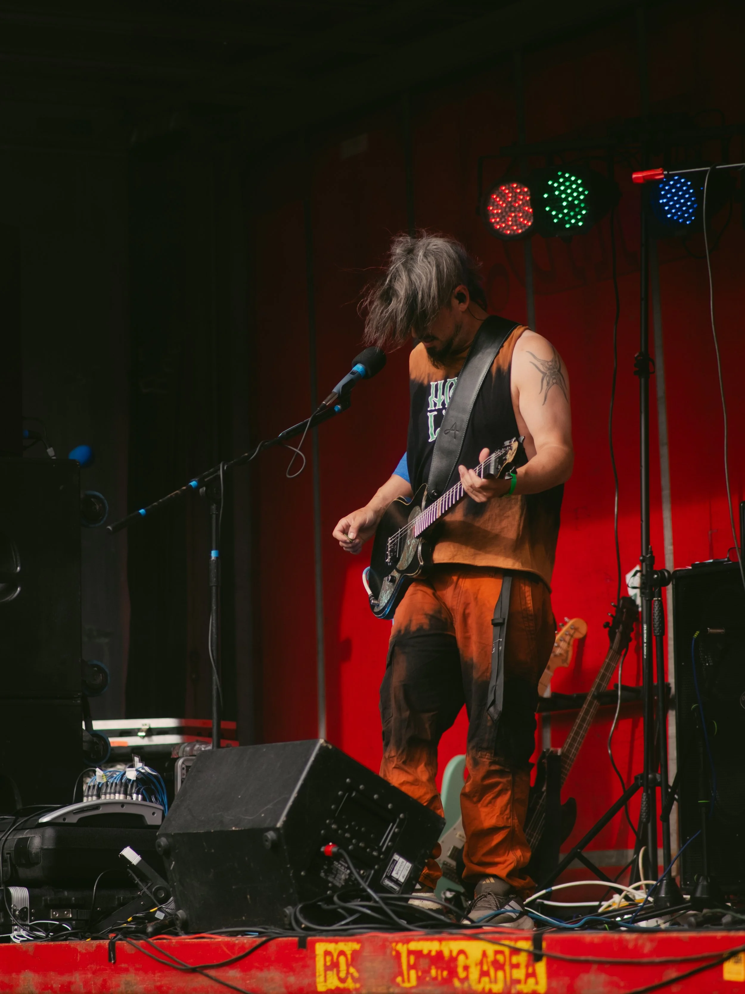 Musician with long hair playing an electric guitar on stage, with red and blue stage lights above.
